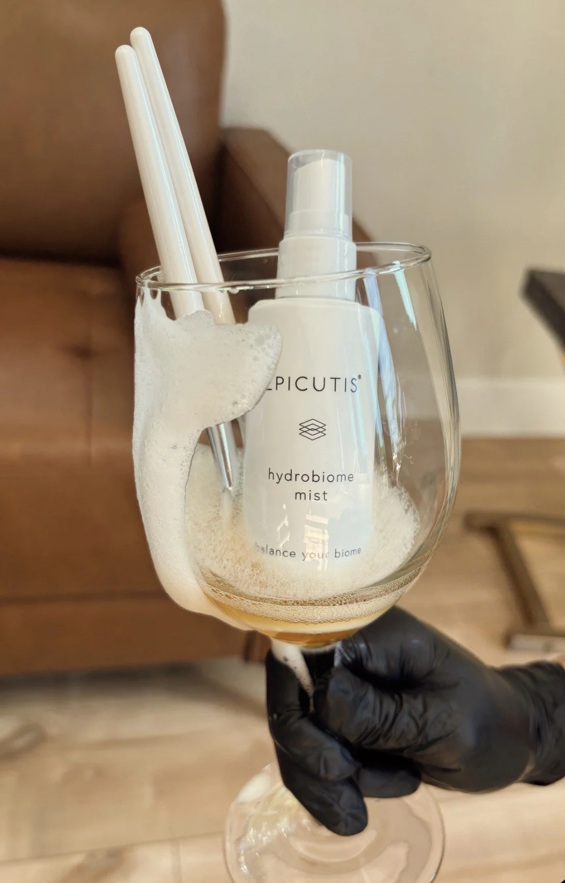 A hand wearing a black glove holding a wine glass filled with foam and a white spray bottle inside, with straws and a spray nozzle sticking out of the foam. The foam clings to the inside of the glass and the bottle, which has the words 'PICUTIS hydobiome mist' printed on it.
