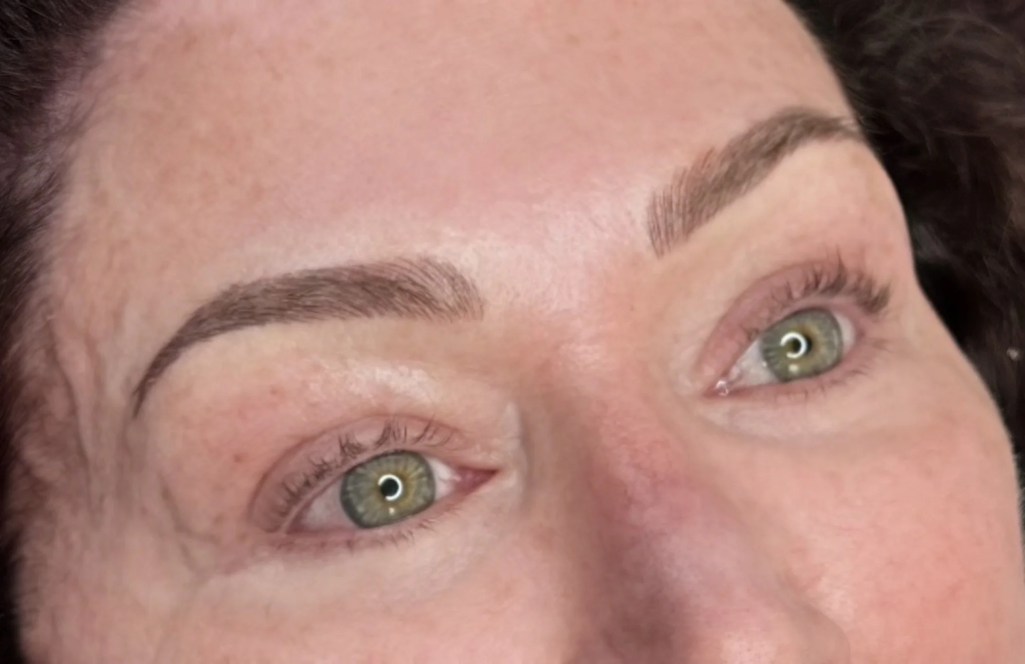 Close-up of a woman's face focusing on her eyes and eyebrows. She has light skin, green eyes, natural eyebrow shape, and makeup-free skin.
