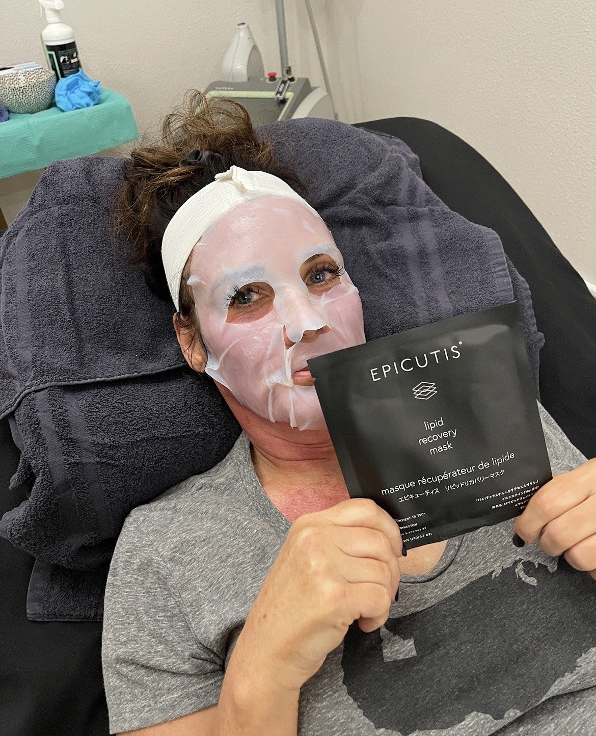 Woman lying on a bed with a gray facial sheet mask, holding a black package labeled 'Epicutis lipid recovery mask'.