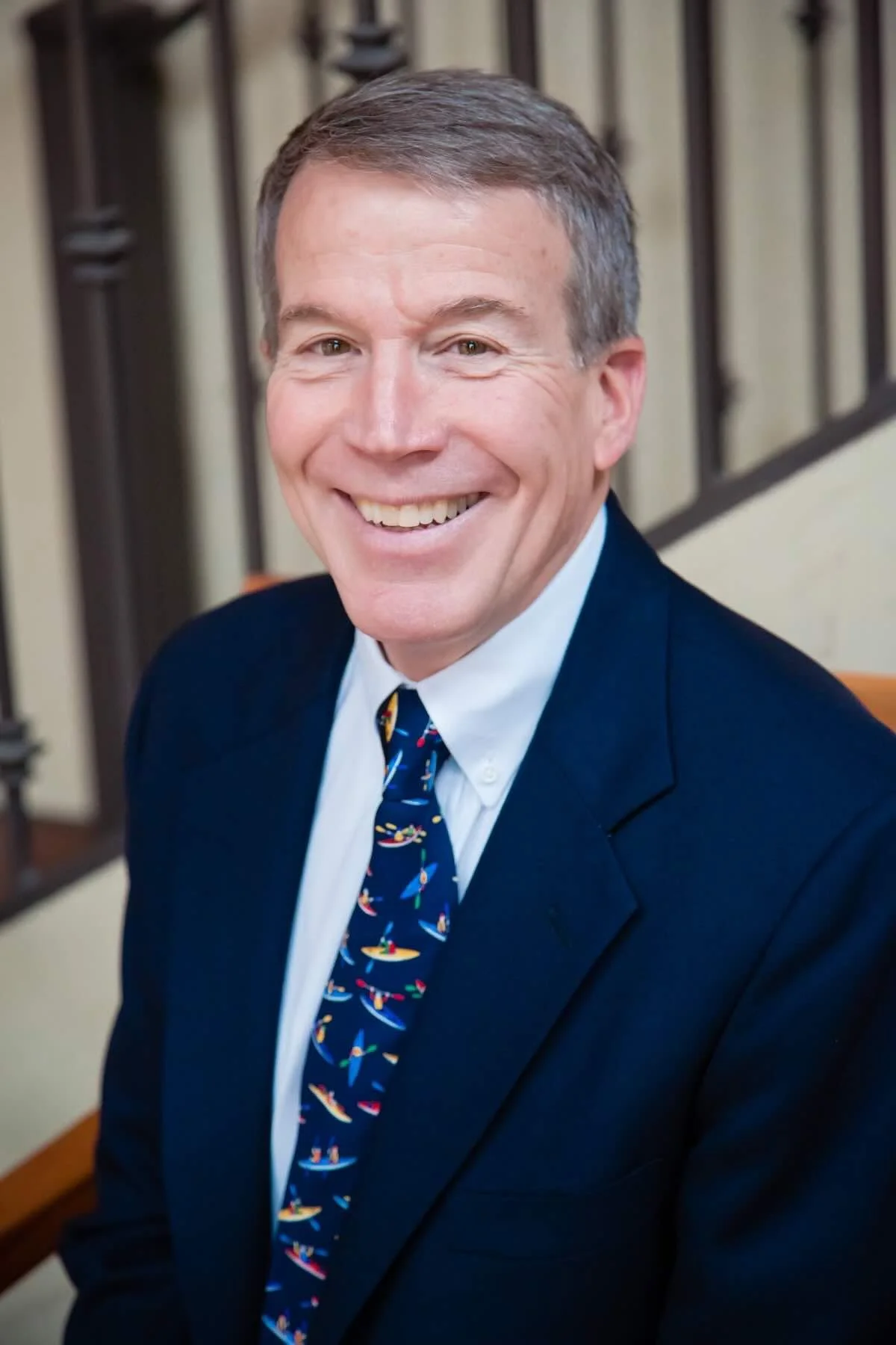 A smiling man with short gray hair, wearing a navy blazer, white dress shirt, and a colorful tie with sailboats, sitting on a bench with a railing in the background.