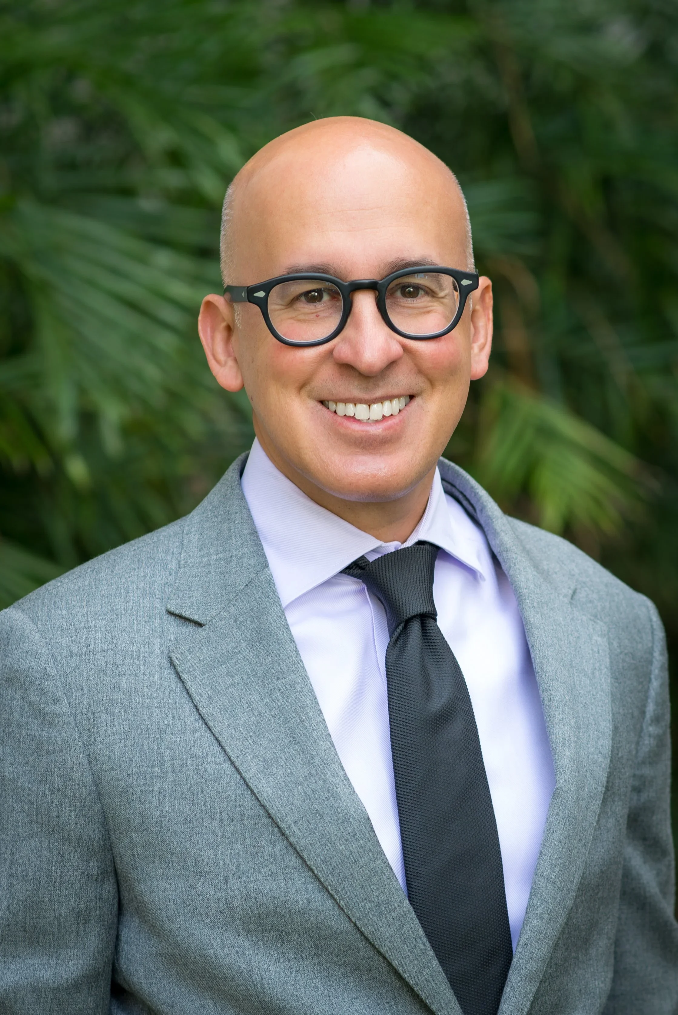 Professional man with glasses smiling outdoors, wearing a gray suit, white shirt, and black tie, with green foliage in background.