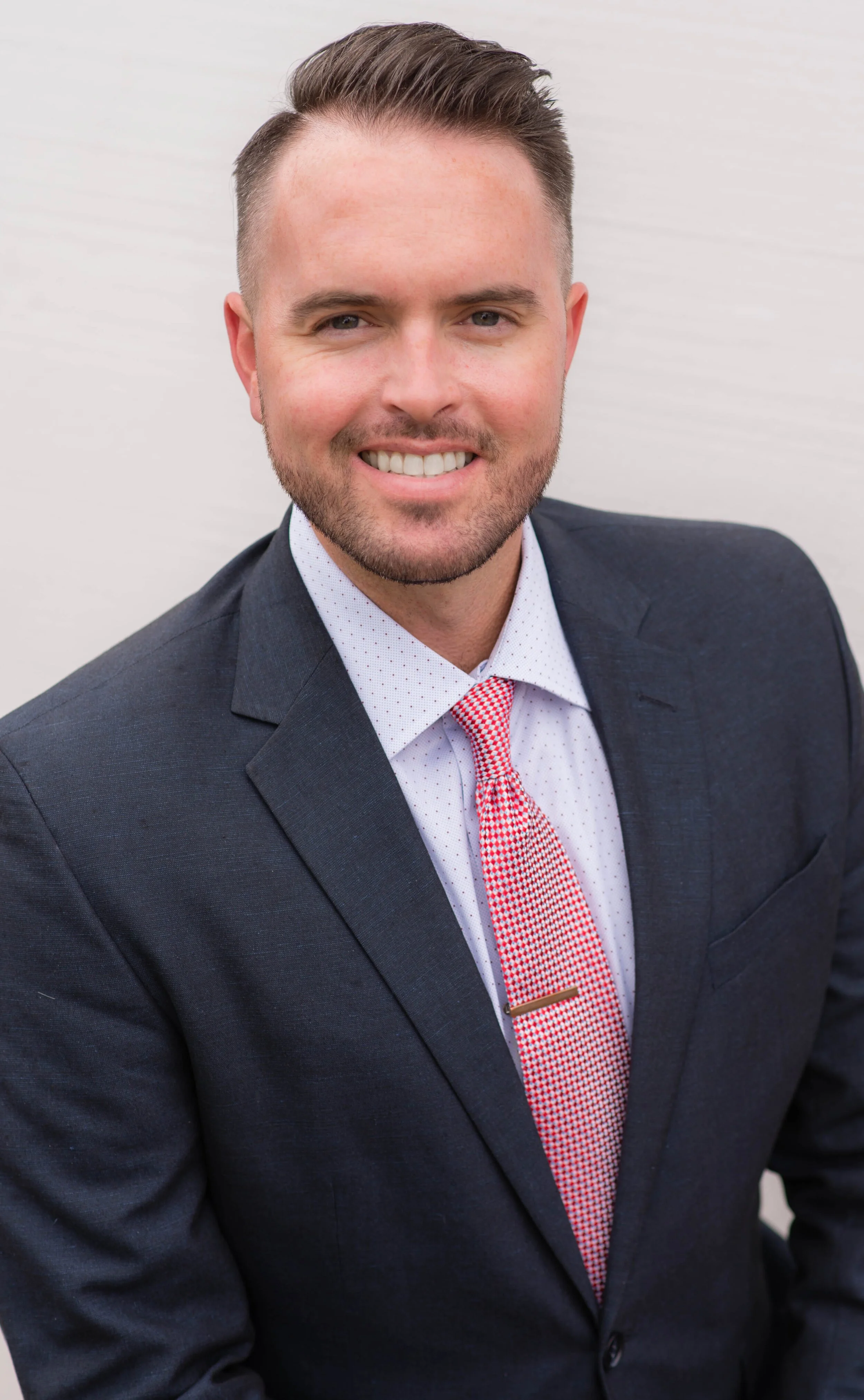 Headshot of a smiling man in a dark suit, white shirt with small polka dots, red and white checkered tie, against a plain light background.