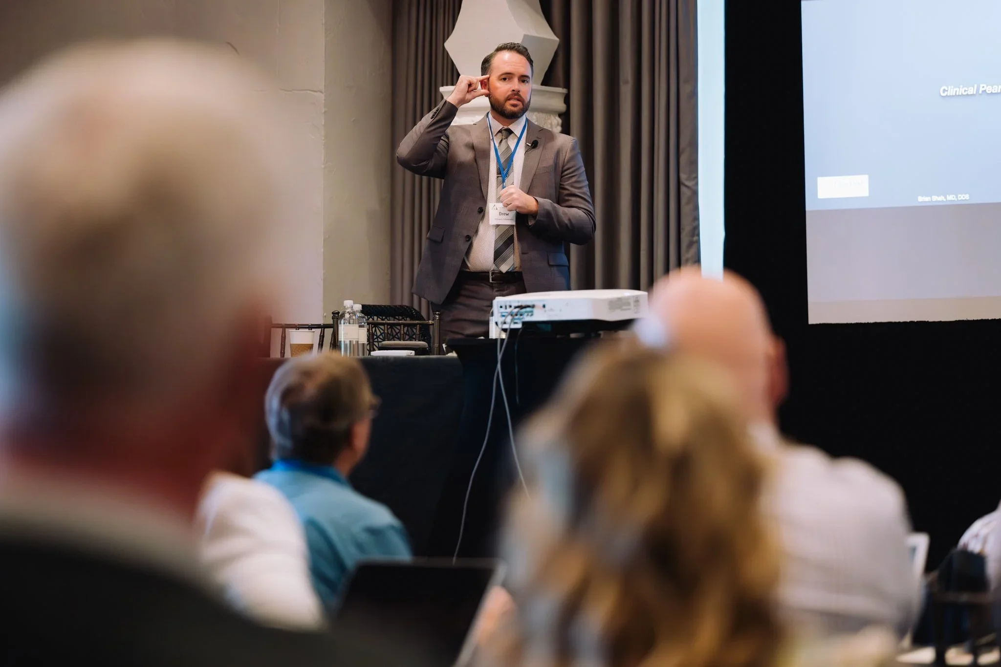 A man in a grey suit giving a presentation at a conference, with a slide projected on a screen and audience members seated in front of him.