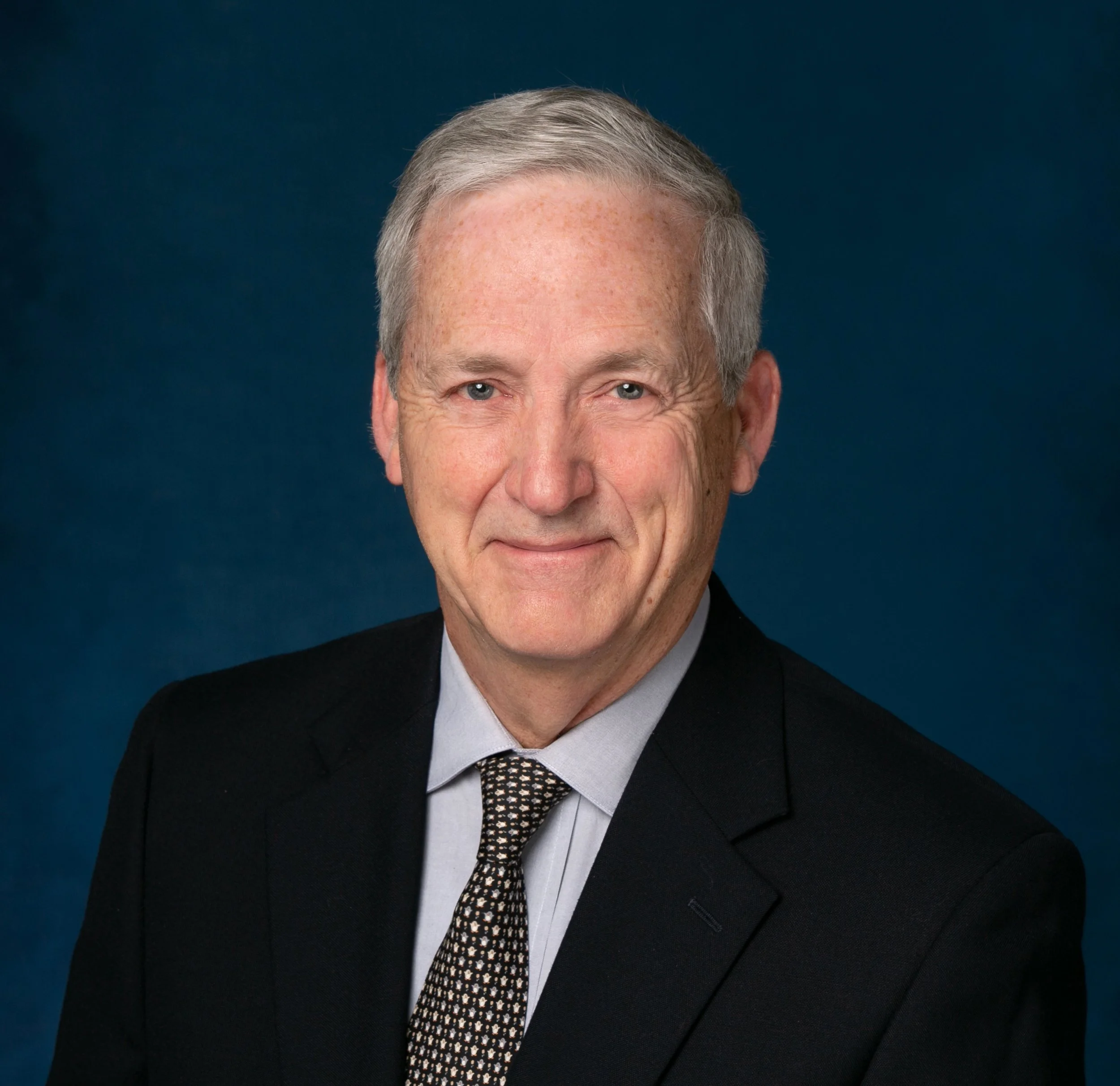 A professional headshot of an older man with gray hair, wearing a dark suit, light shirt, and patterned tie, against a dark blue background.