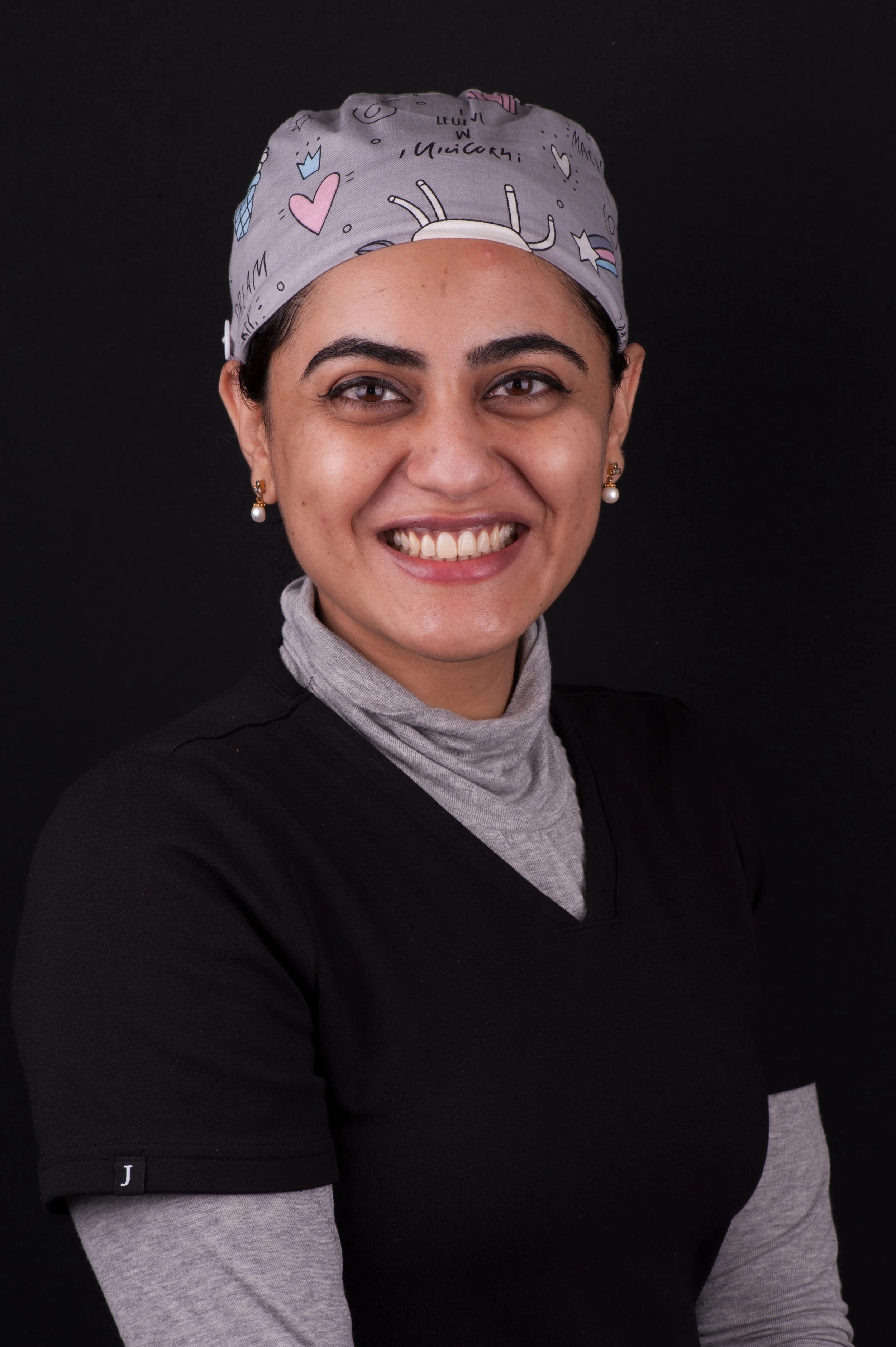 A woman smiling, wearing a gray medical cap with colorful designs and writing, pearl earrings, a gray turtleneck, and a black shirt against a black background.