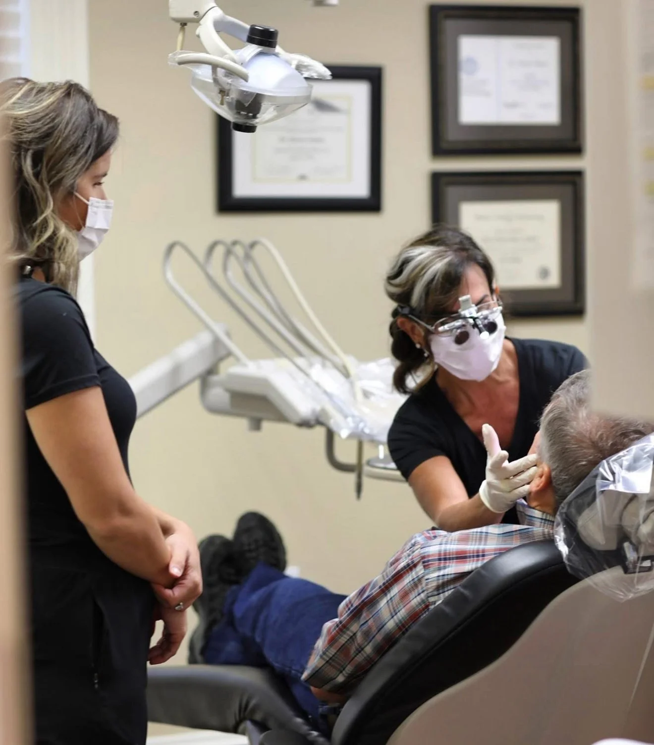 Dentist examining an elderly patient in a dental office, with a dental assistant and another staff member present, all wearing masks.
