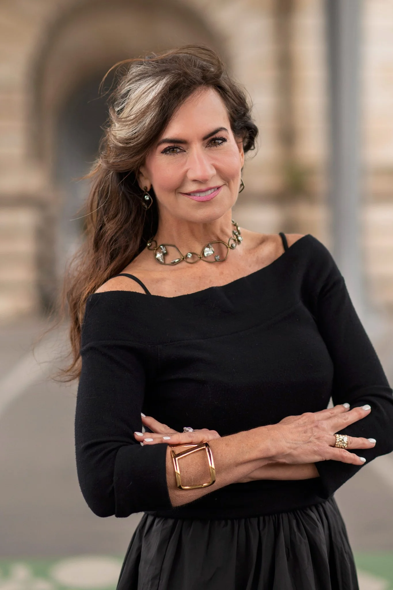 A woman with wavy brown hair and light skin, wearing a black off-shoulder top and signature jewelry, is posing with arms crossed in front of a blurred brick building background.