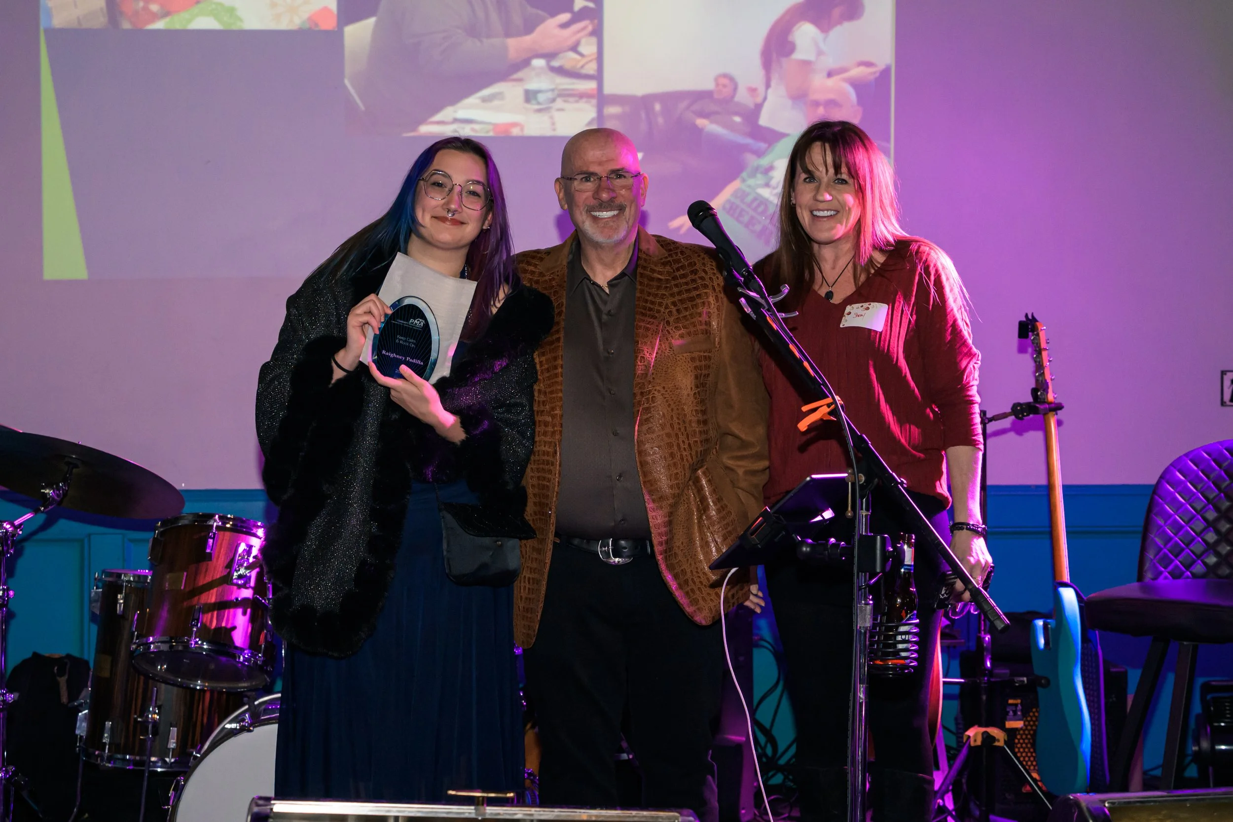 Three people on stage at an awards event, with a drum set in the background. The woman on the left is holding a plaque, the man in the middle is smiling, and the woman on the right is smiling near a microphone stand.