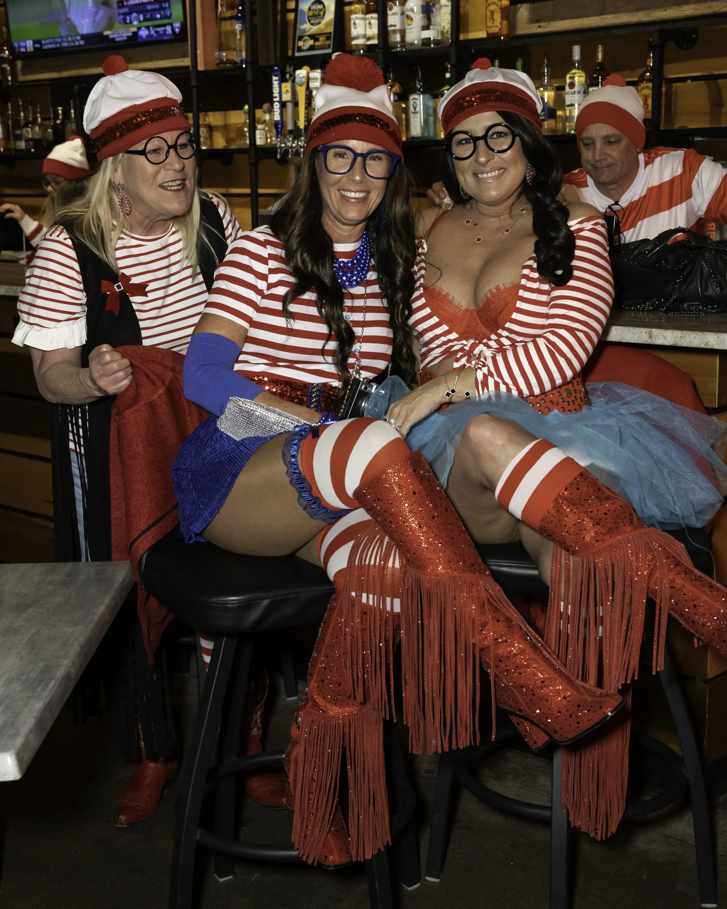 Four people at a bar dressed in festive, Waldo-themed costumes. Three women are seated in the foreground, and a man is in the background. The women are wearing striped red and white tops, Santa hats, and colorful accessories. 