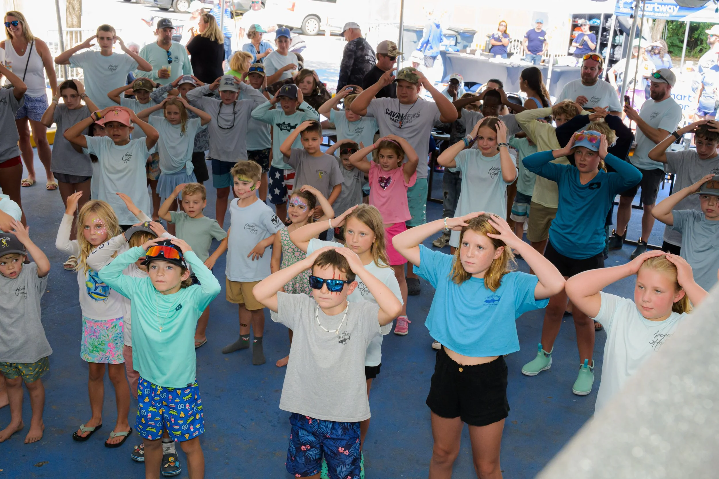 A group of children and some adults gathered outdoors under a tent, participating in a group activity at Ballyhoos, with many children placing their hands on their heads.