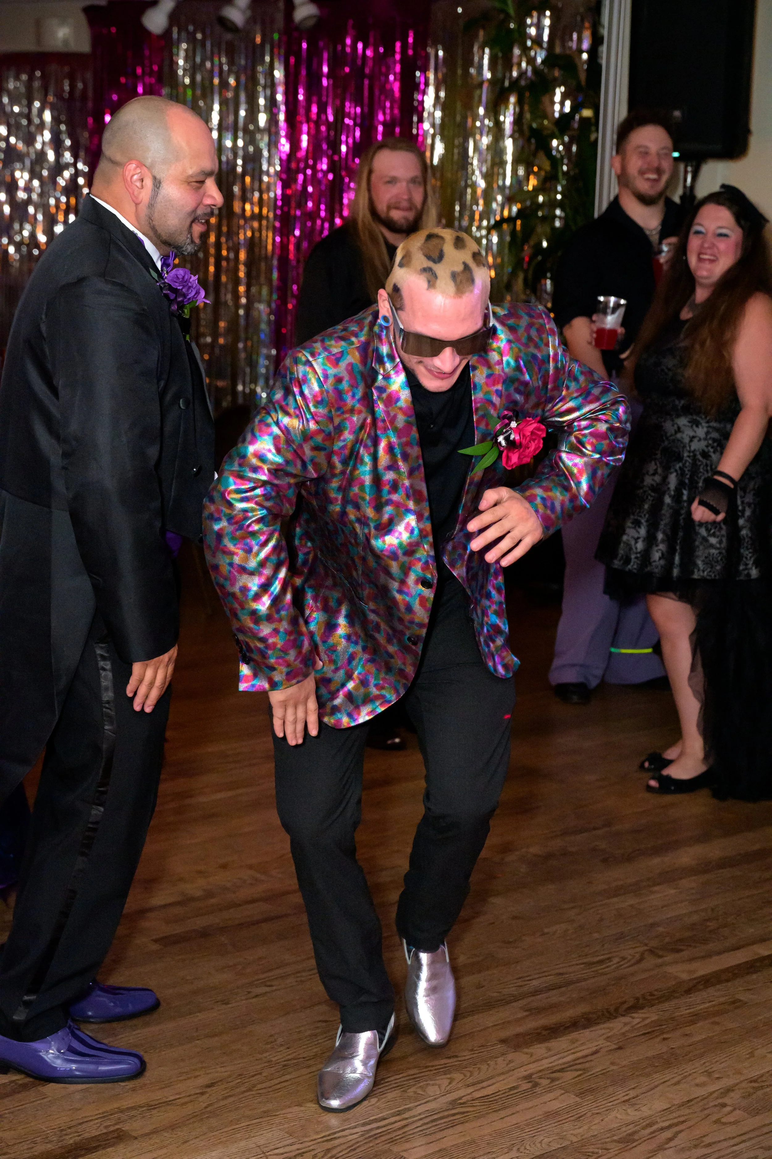 Person dancing in colorful jacket, metallic shoes, with large sunglasses at party with people in background.