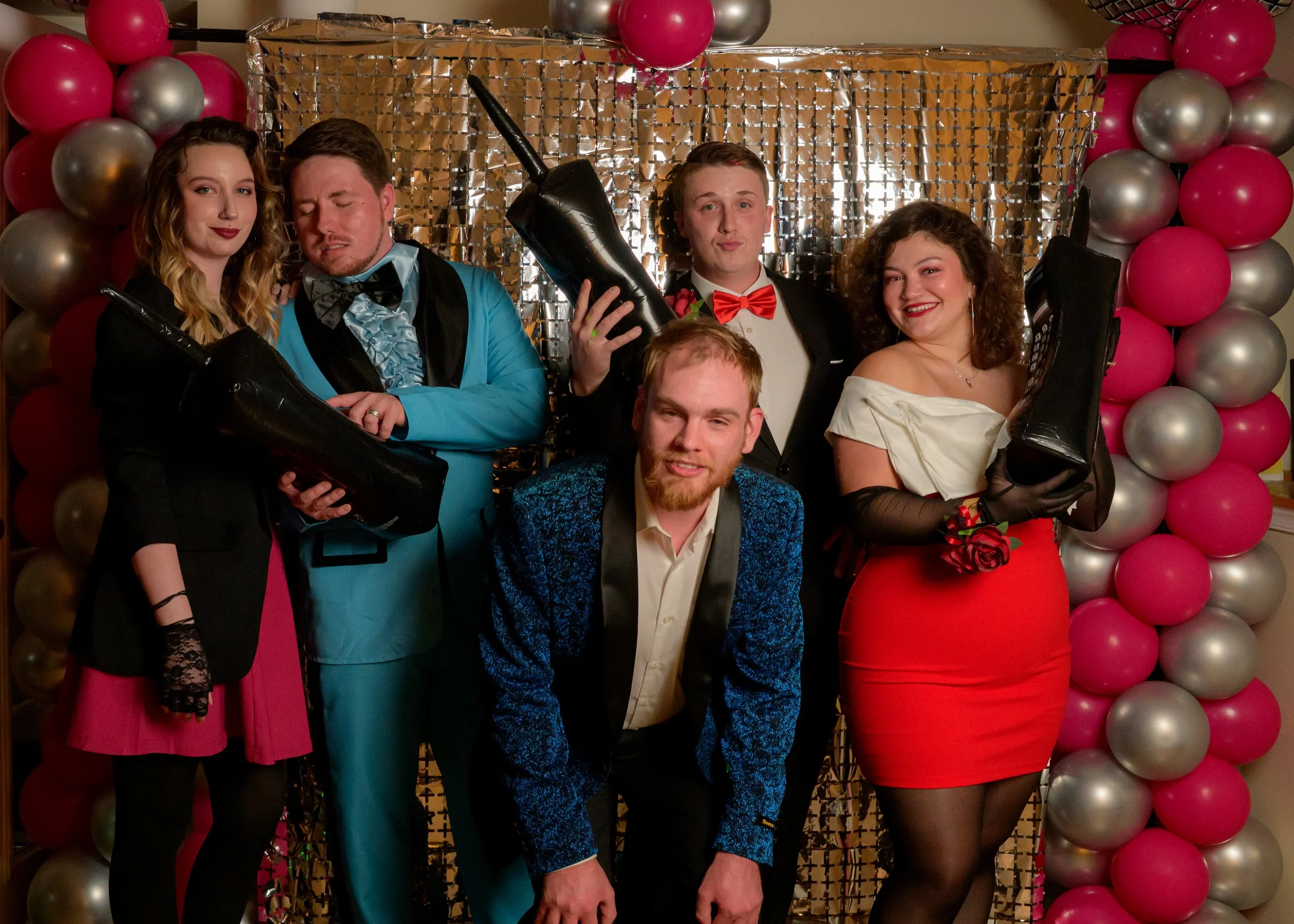 Group of six young adults dressed in formal attire posing at a party with pink and silver balloon decorations and a golden backdrop.