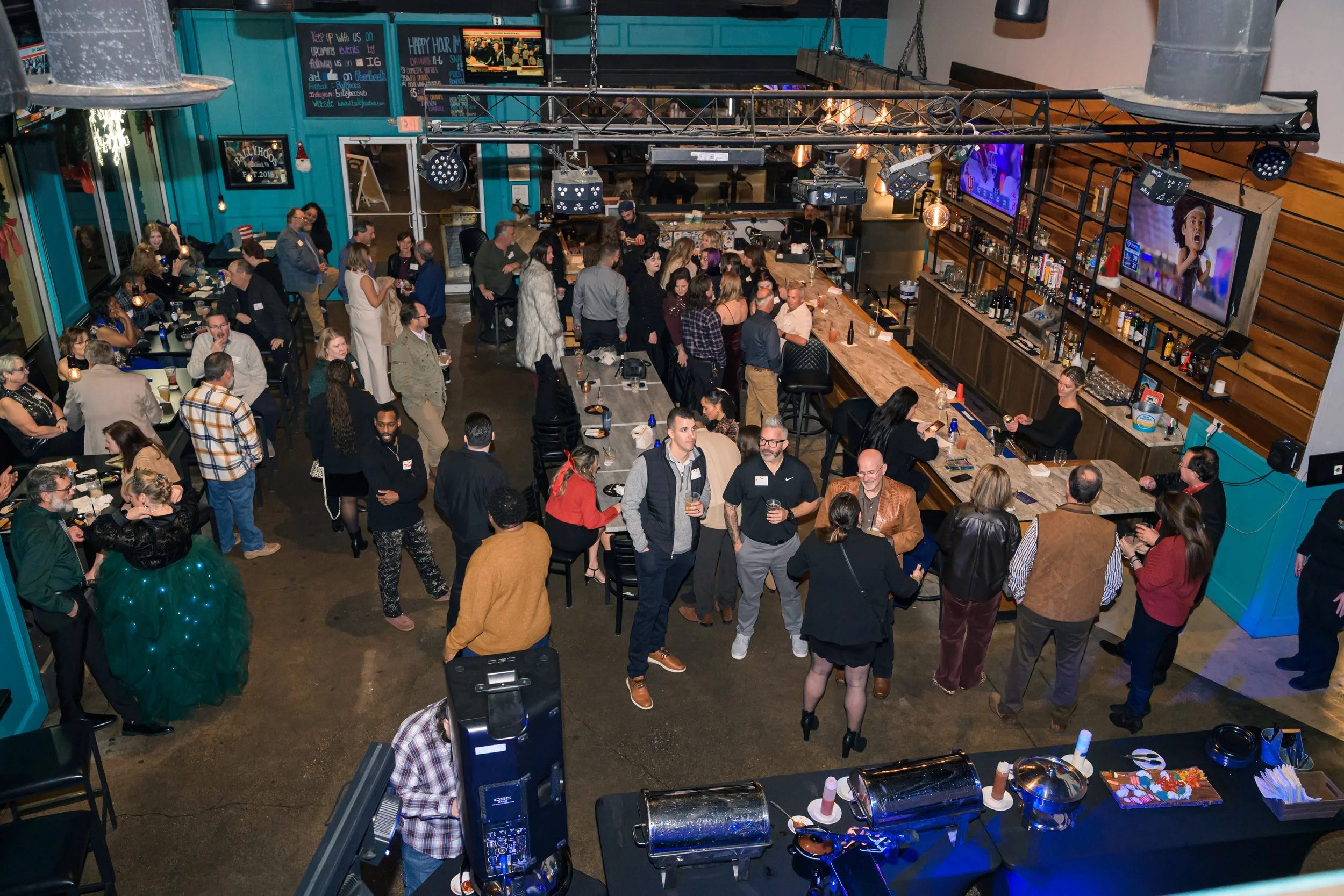 A lively bar scene with numerous people socializing, some standing and some seated at tables. The bar area has a wooden countertop and shelves stocked with bottles. Large television screens display various images, and the space features a teal and wo