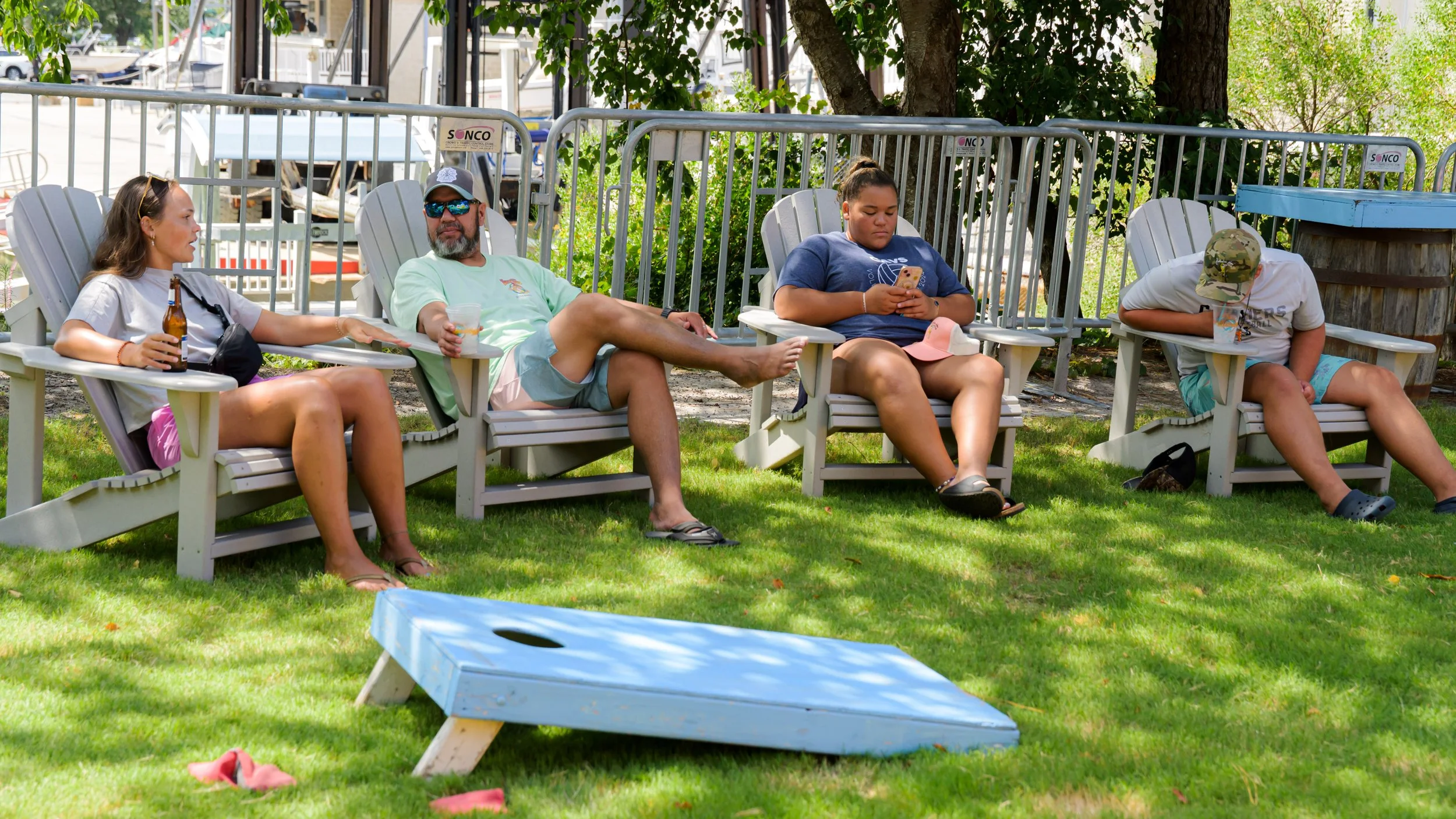 Four people sitting on Adirondack chairs outdoors, two men and two women, with one woman holding a beer bottle, one man with a drink, a woman looking at her phone, and a man with a cap and hydration bottle. There is a cornhole game nearby on a grassy