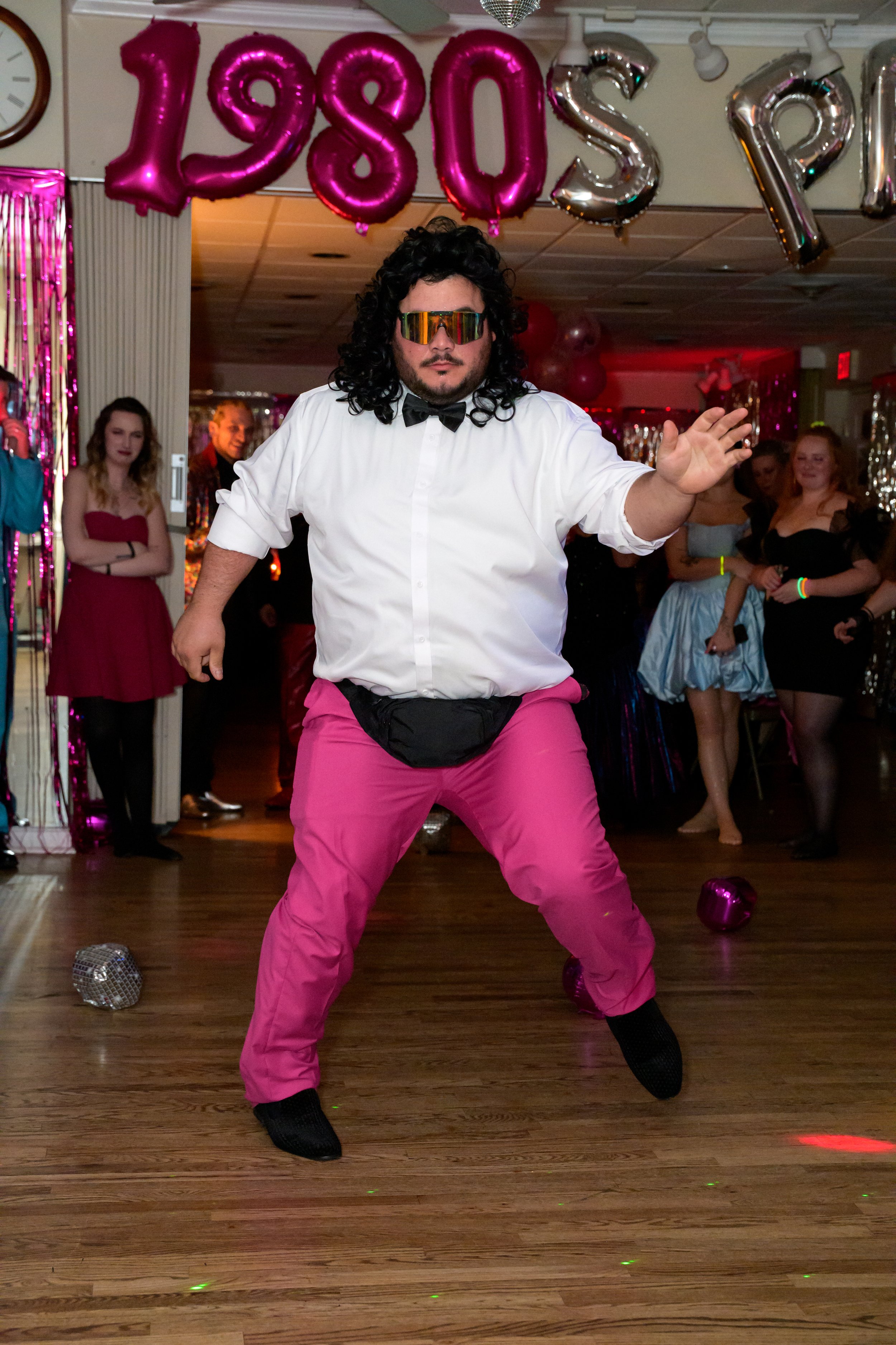 Man dancing at 1980s themed party, wearing pink pants, white shirt, black fanny pack, black shoes, and sunglasses, with decorative balloons spelling '1980s' in the background and a group of party guests watching.