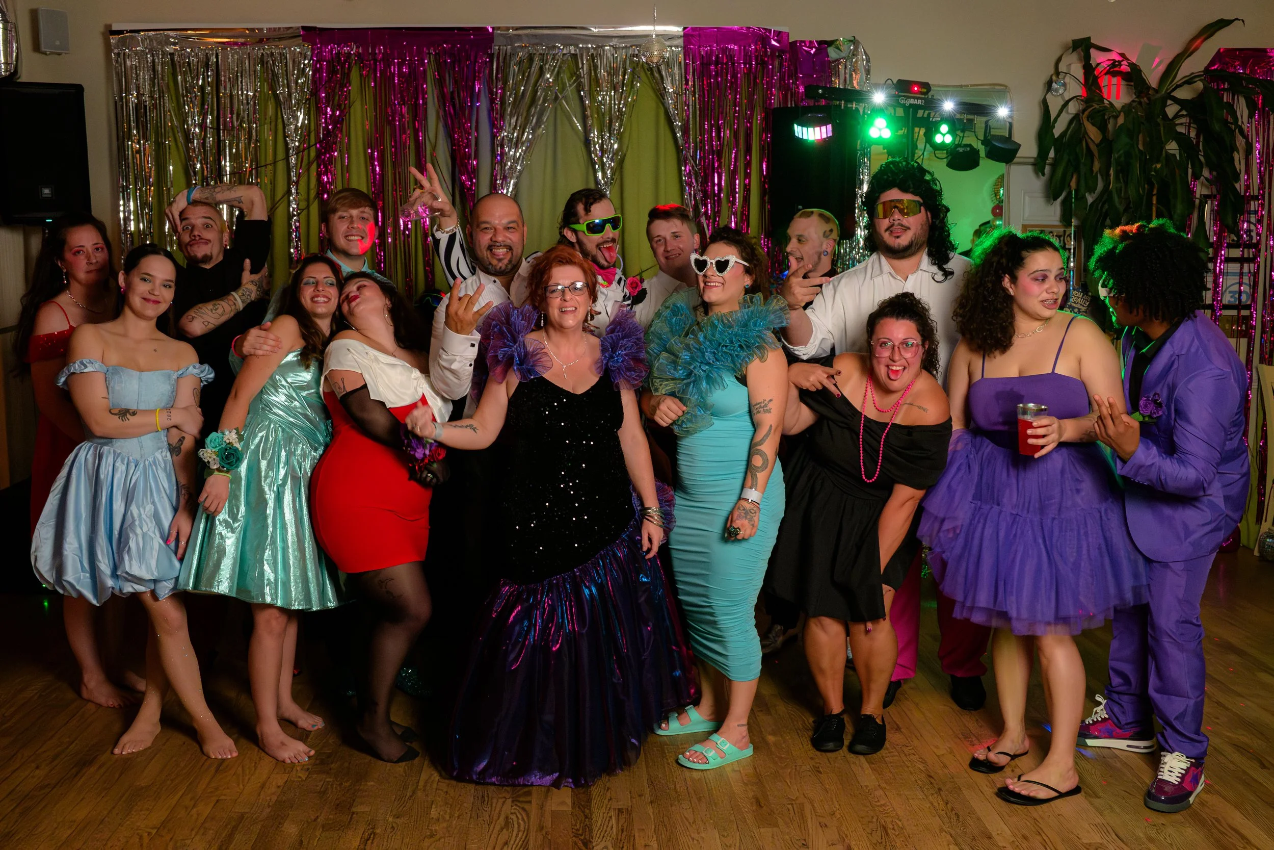 Group of people dressed in colorful costumes at a party, standing in front of shiny decorations and a green light on stage.