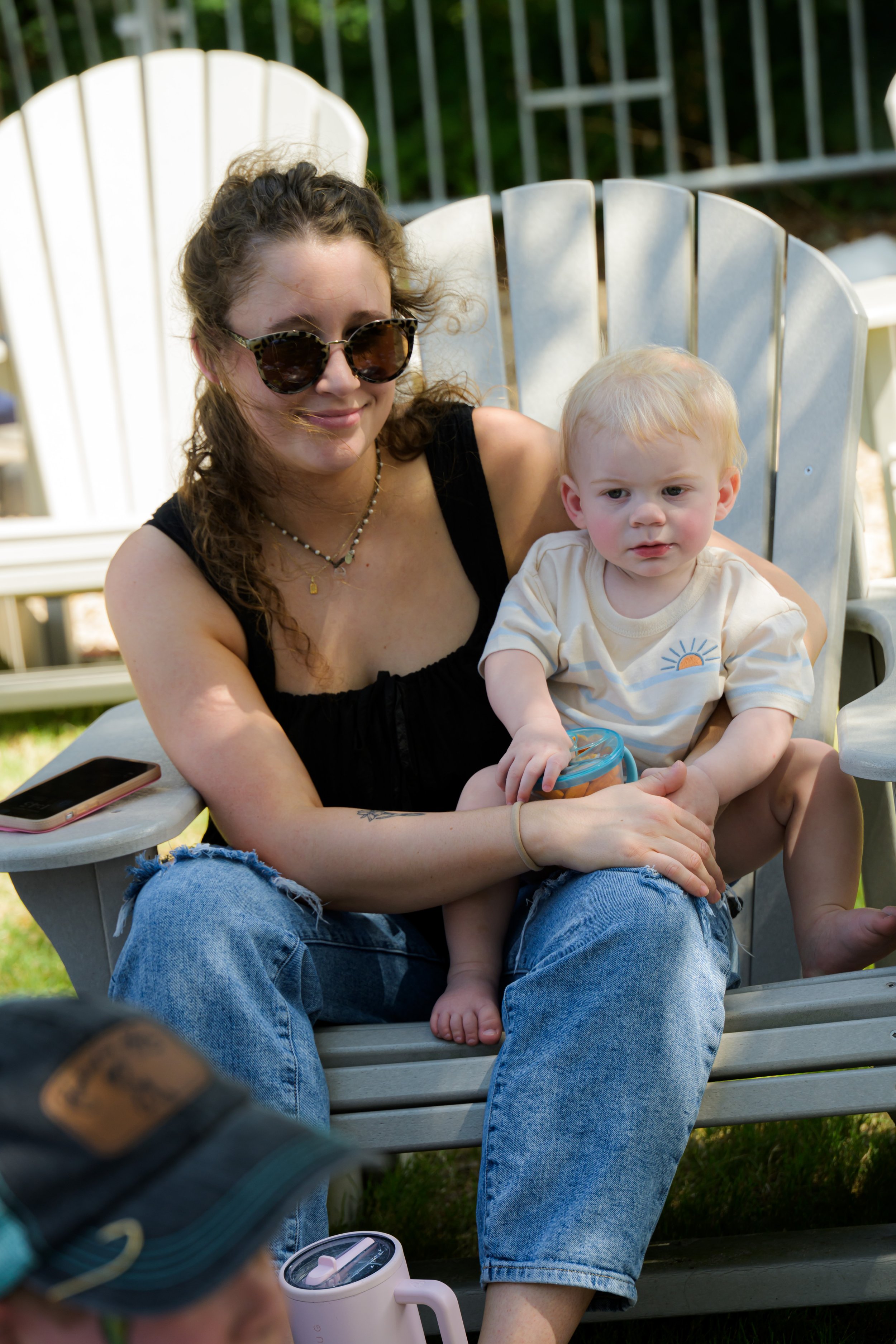 A woman with curly hair wearing sunglasses and a black tank top, sitting on a white Adirondack chair outdoors, holding a young blond boy in her lap. The boy is wearing a cream T-shirt with a sun graphic and is holding a small toy.