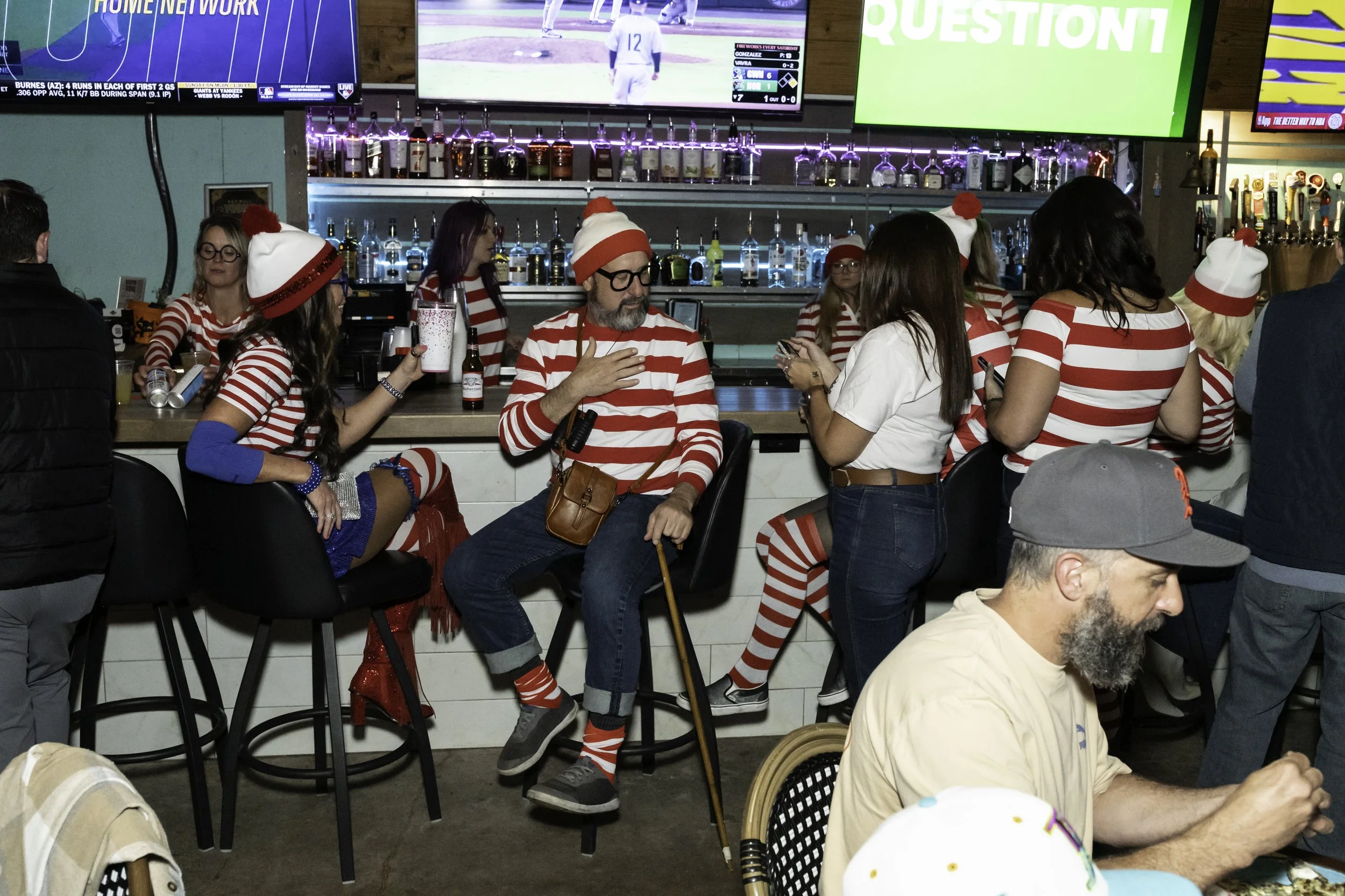People dressed in festive red and white striped outfits like Waldo, gathered at Ballyhoos with TV screens showing sports, and a wall of liquor bottles behind the bar.