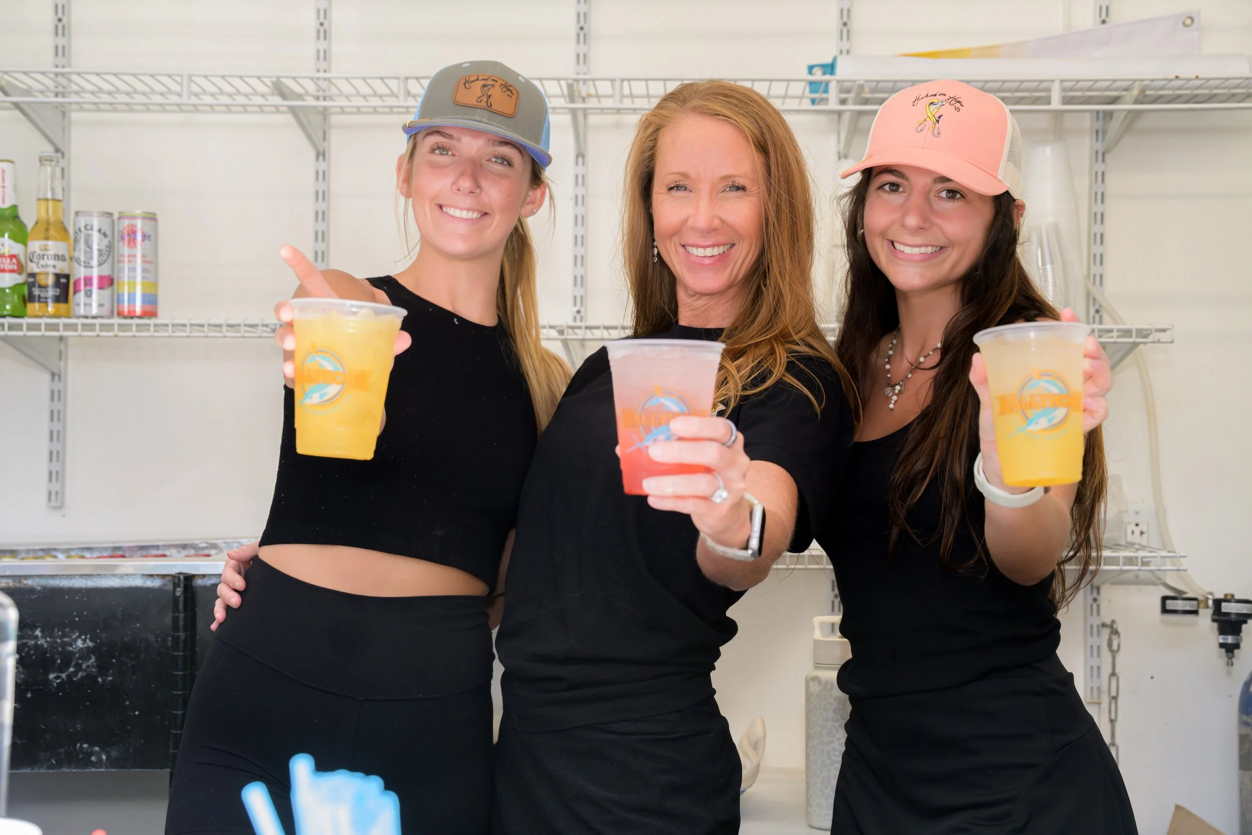 Three women standing behind a bar, smiling, holding colorful drinks in plastic cups, with beer bottles and other bar supplies on shelves in the background.