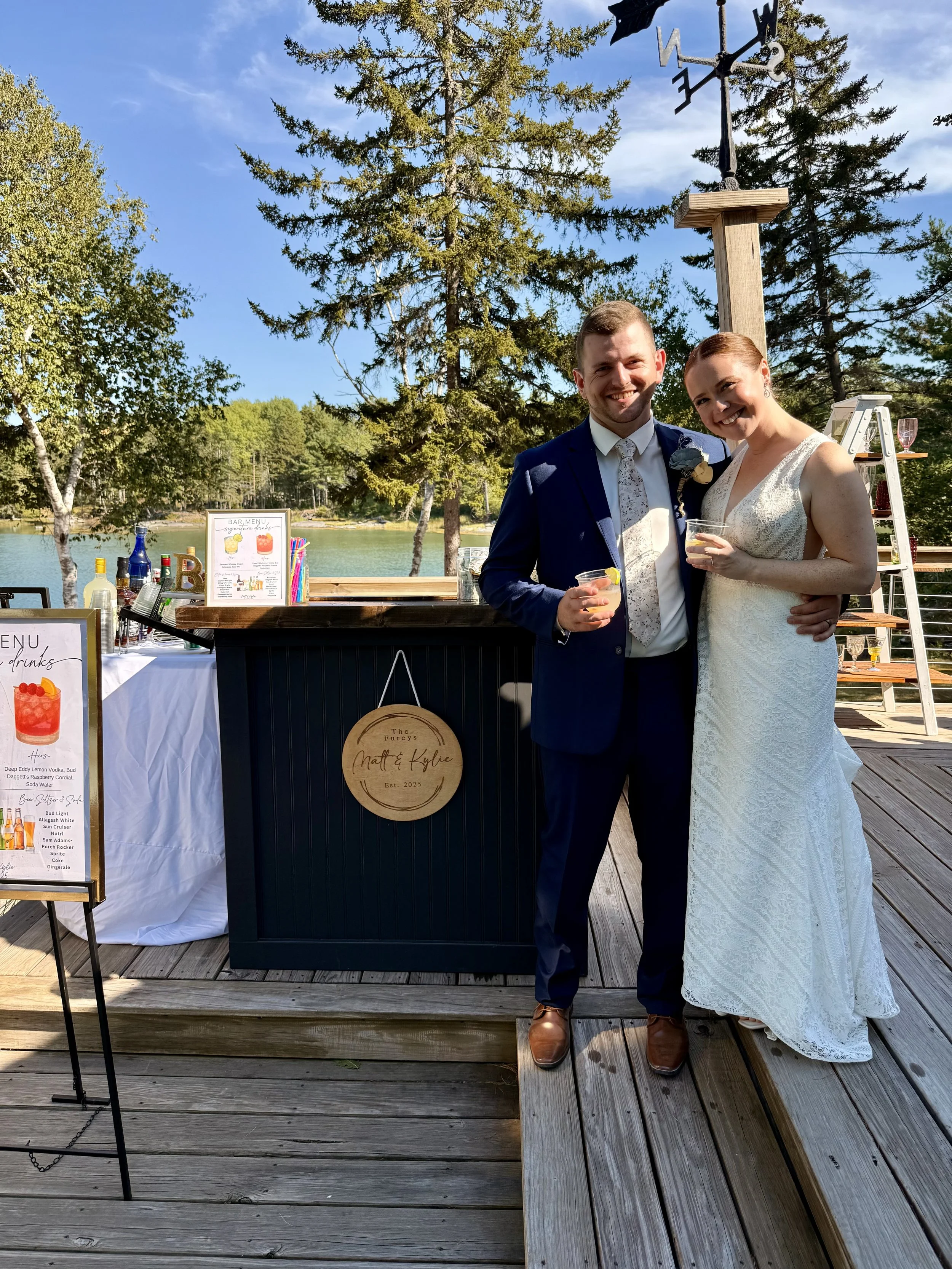 A newlywed couple in wedding attire standing on a wooden deck, holding drinks, smiling, near a bar with a lakeside background and trees.