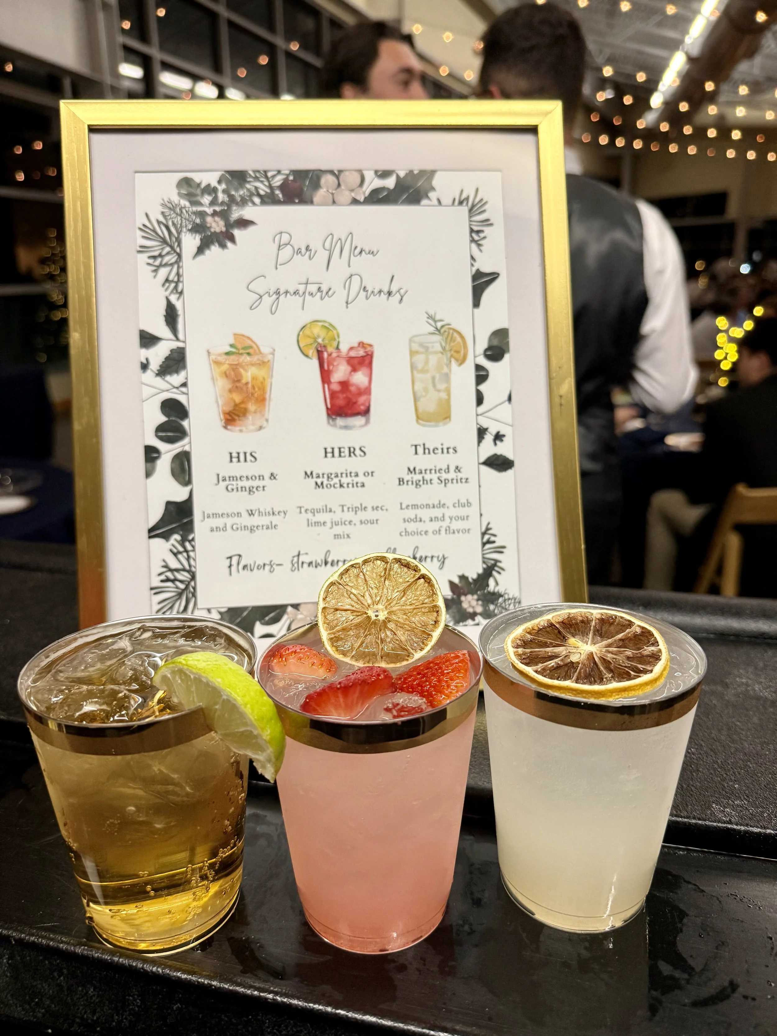 Three cocktails with fruit garnishes on a black serving tray in front of a framed menu at a restaurant. The menu offers three signature drinks: His with Jameson whiskey, Her with tequila or margarita, and Theirs with lemonade or club soda. The drinks