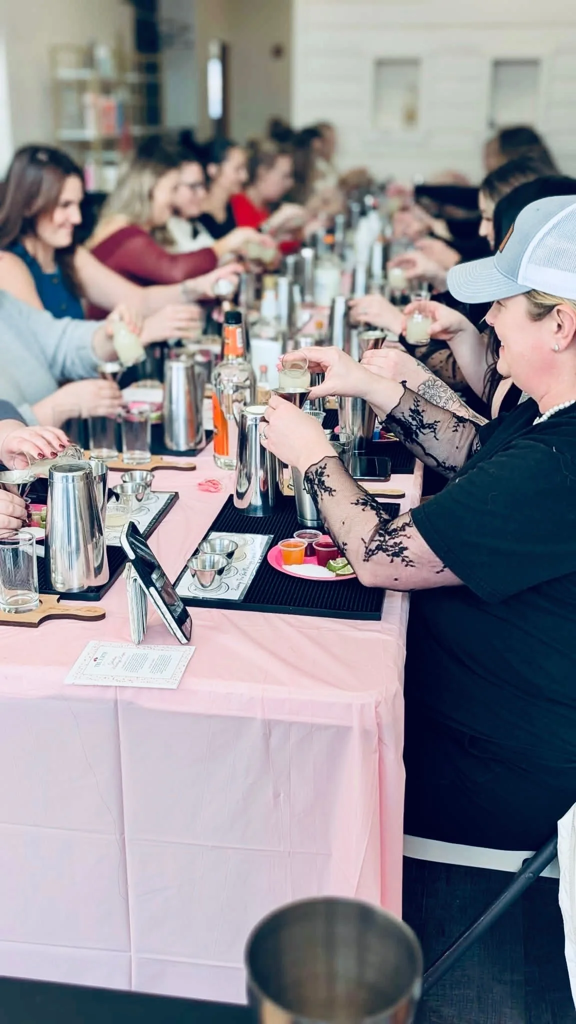 A group of women participating in a guided cocktail tasting or mixology class at a long table with pink tablecloth, various drinks, ingredients, and bartending tools.