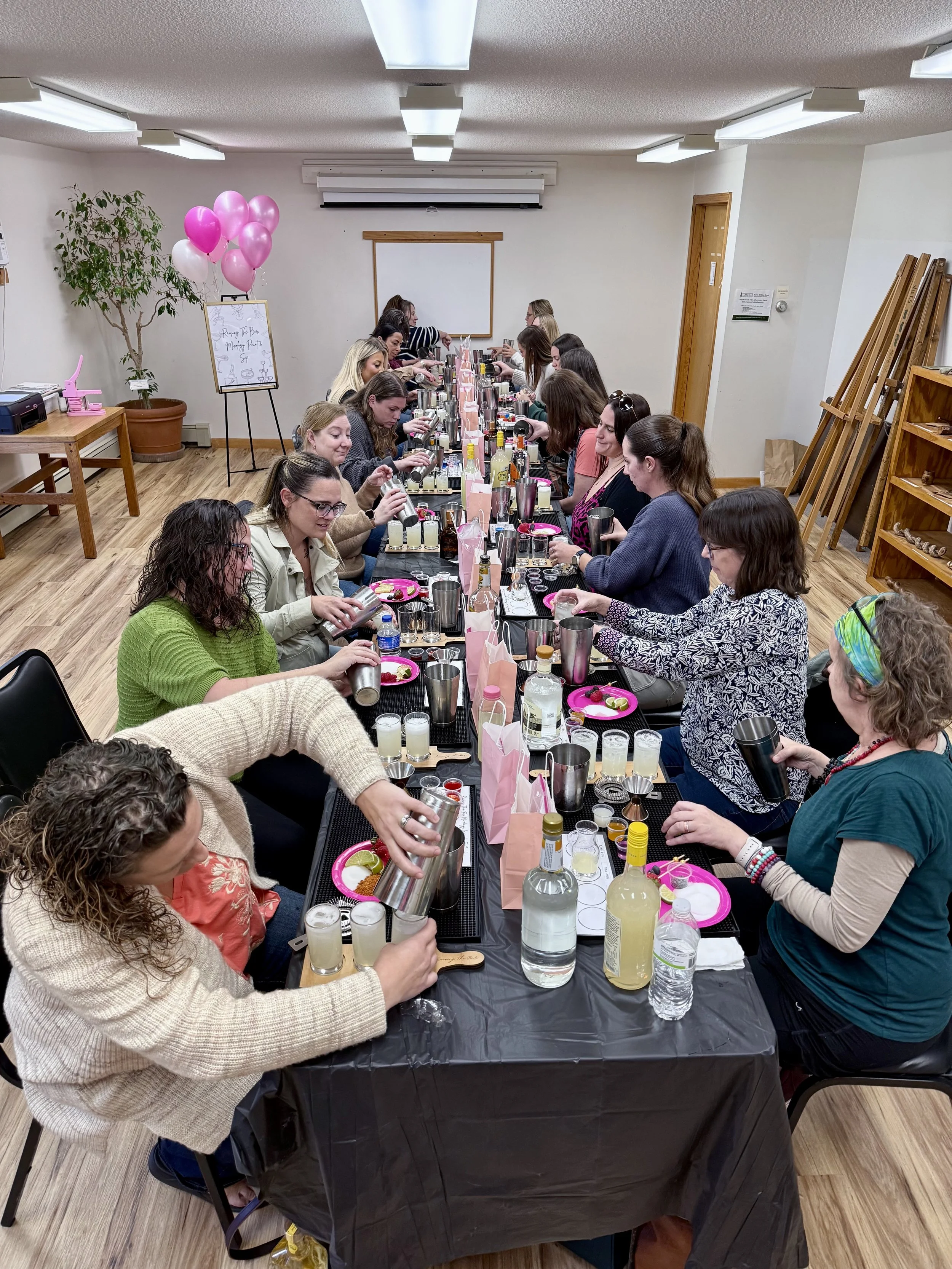 Group of women participating in a makeup or beauty workshop, seated at a long table with cosmetics and drinks in a classroom setting, decorated with pink balloons.