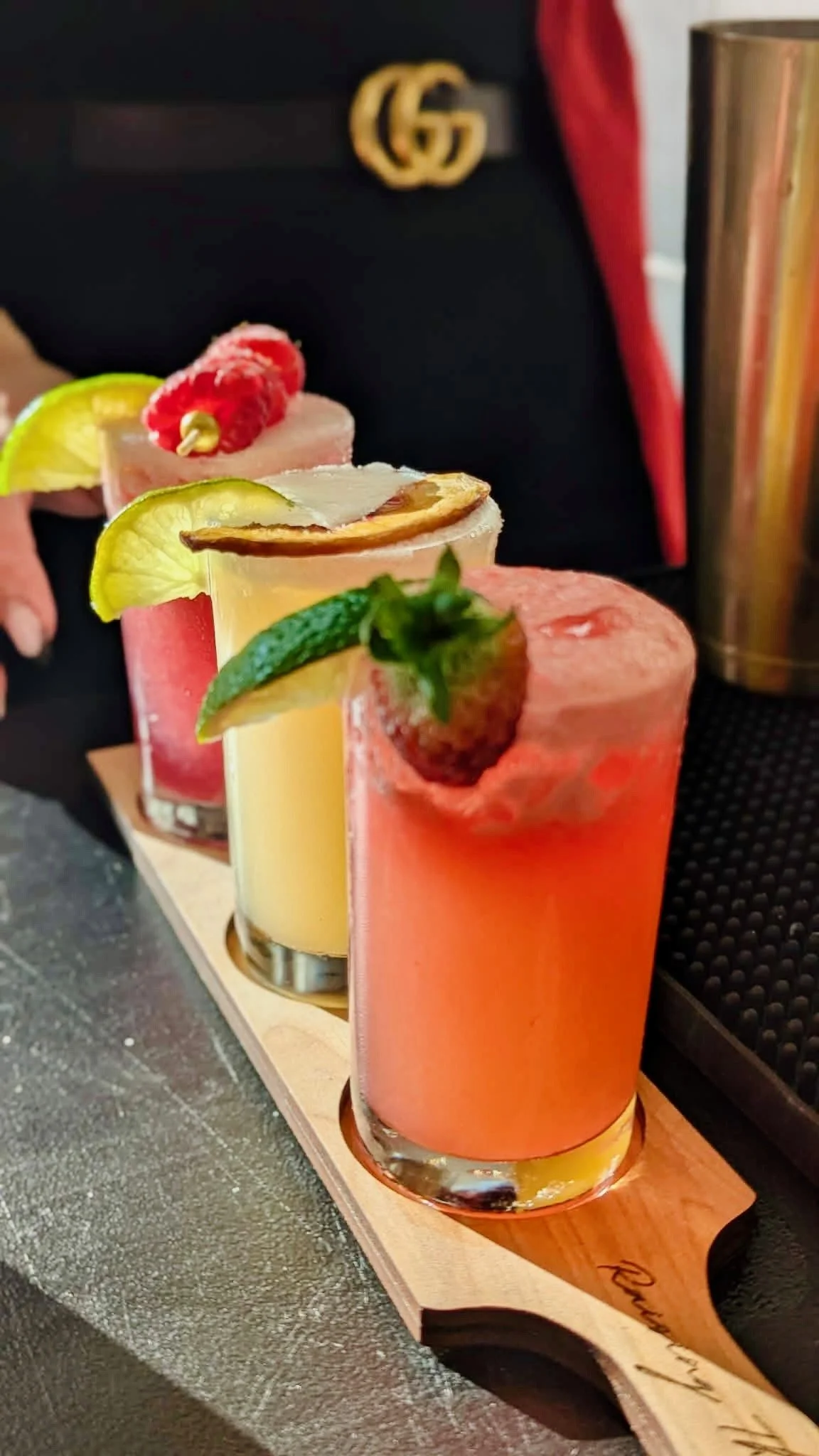 A wooden flight paddle holding three colorful cocktails garnished with fruit slices and berries.