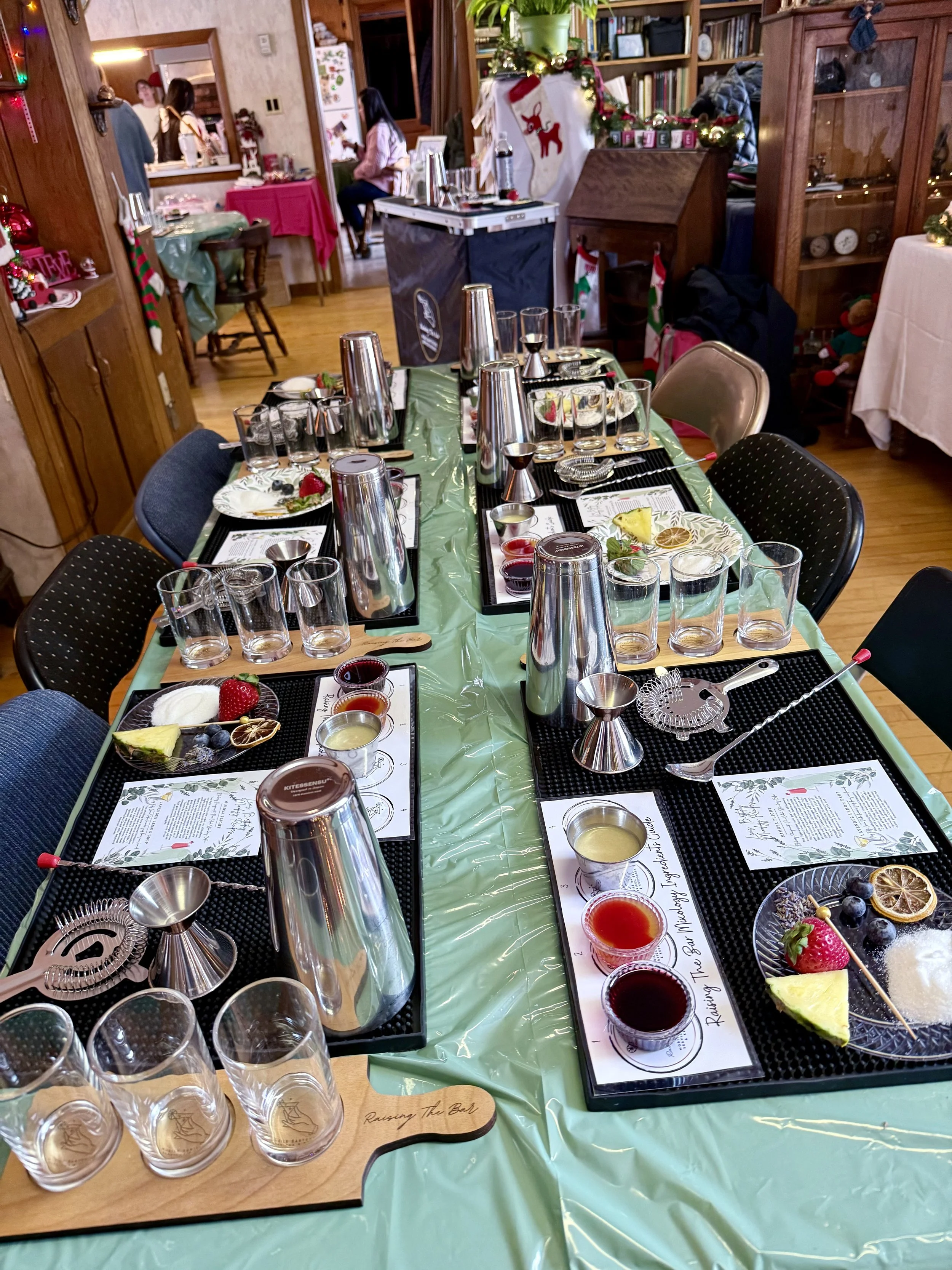 A festive holiday cocktail tasting setup on a long table with black placemats, glasses, and small plates with fruits and sauces, surrounded by chairs in a decorated room with Christmas decorations.