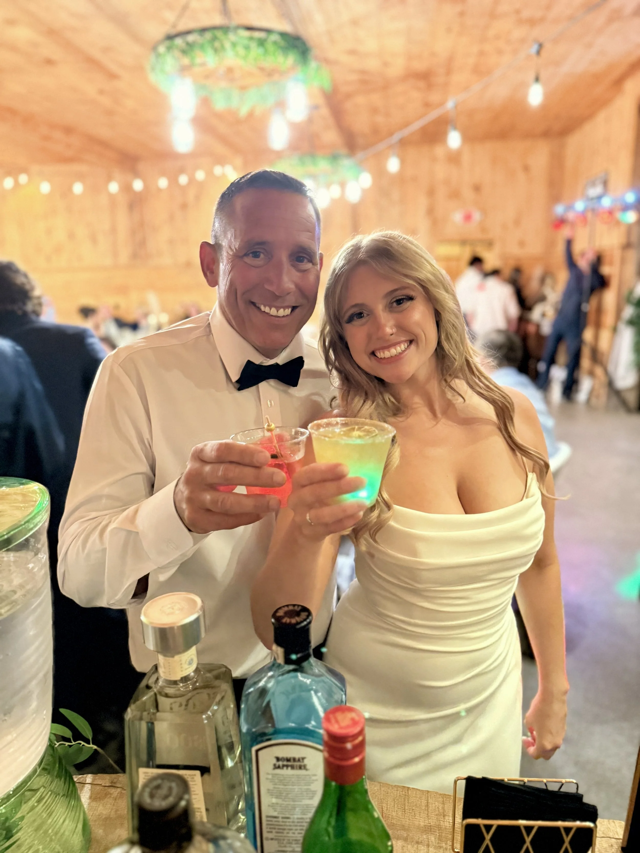 A smiling man in a white shirt and black bowtie and a smiling woman in a white dress are holding colorful drinks, celebrating at an indoor event with wooden walls and string lights in the background.