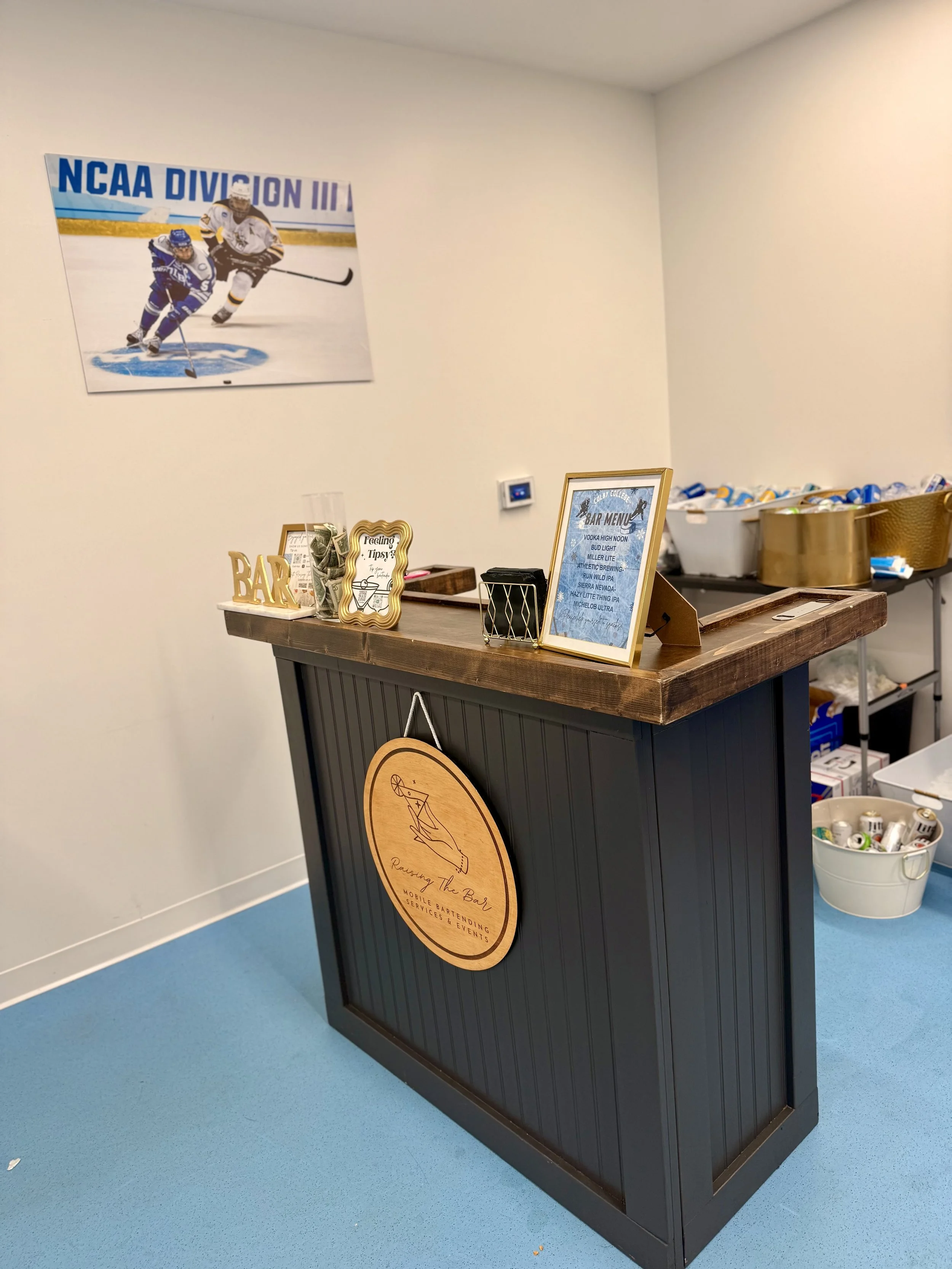 A bar counter with a sign that reads 'Raising The Bar', a framed bar menu, and other small decor items. Behind the counter is a large tub filled with cans, and on the wall, there is a hockey poster of two players skating.