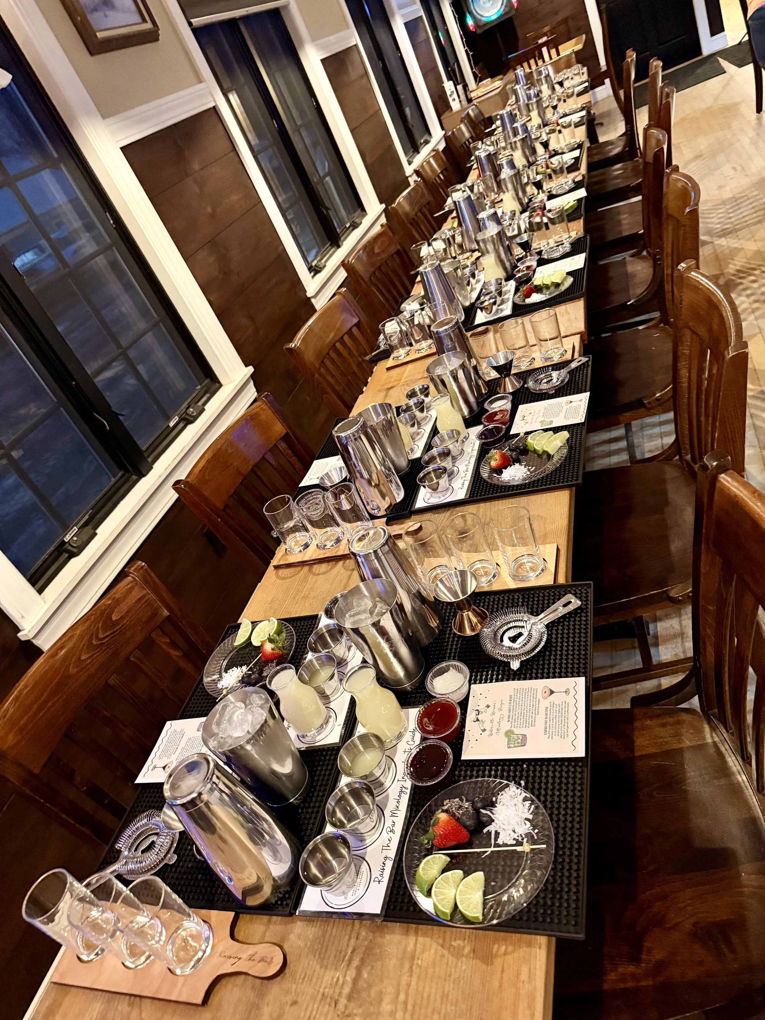 A long wooden table set for a cocktail or drink tasting event, with various shot glasses, cocktail shakers, and ingredients, along with small plates of lime wedges, strawberries, and garnishes.