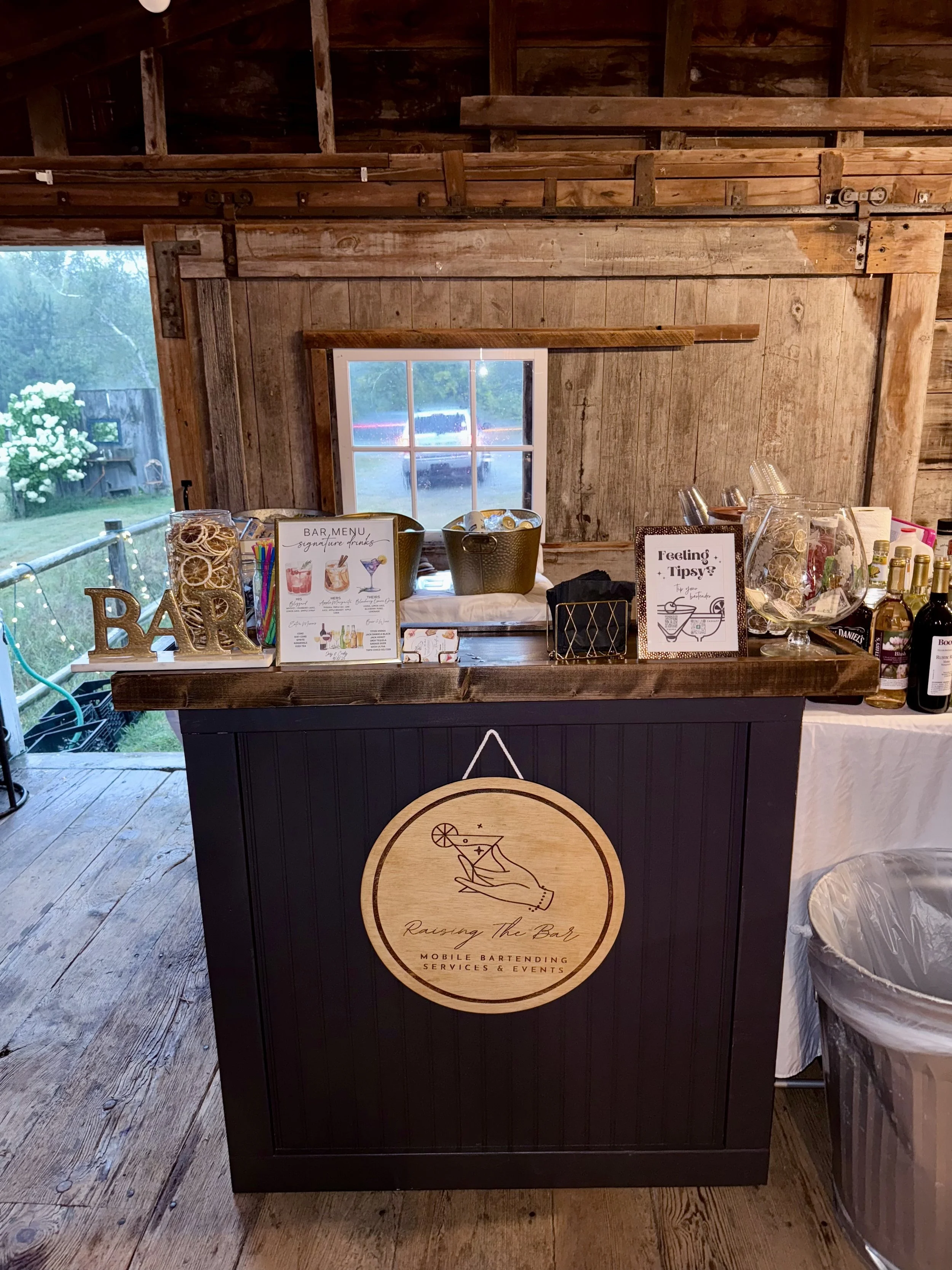 A rustic bar setup inside a wooden barn with a sign that reads "Raising the Bar Mobile Bartending Services & Events" featuring a hand holding a cocktail glass with a lemon wedge. The bar has various drink supplies, a menu, and decorative items, with 