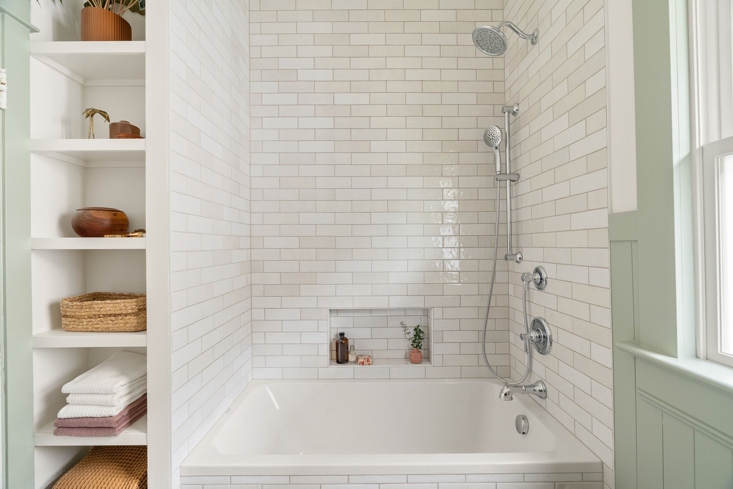 A bathroom shower with two showerheads mounted on white-tiled wall, a built-in niche with toiletries and a small plant, and a window on the right side.