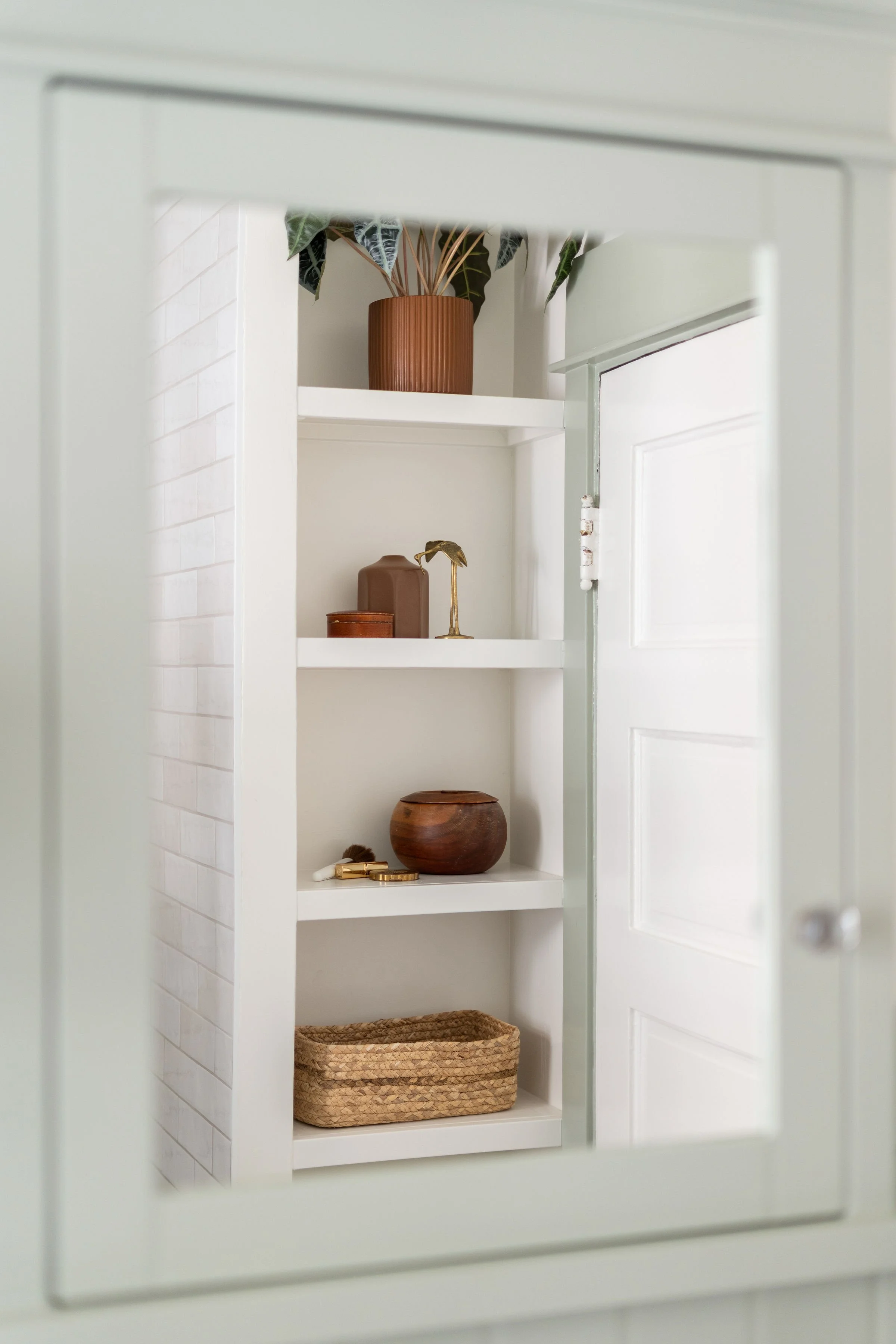 A view through a mirror showing a white built-in shelf with decorative items, including a potted plant, a brown vase, a gold flamingo figurine, a wooden bowl, a basket, and various small objects, in a bright room with white walls and door.