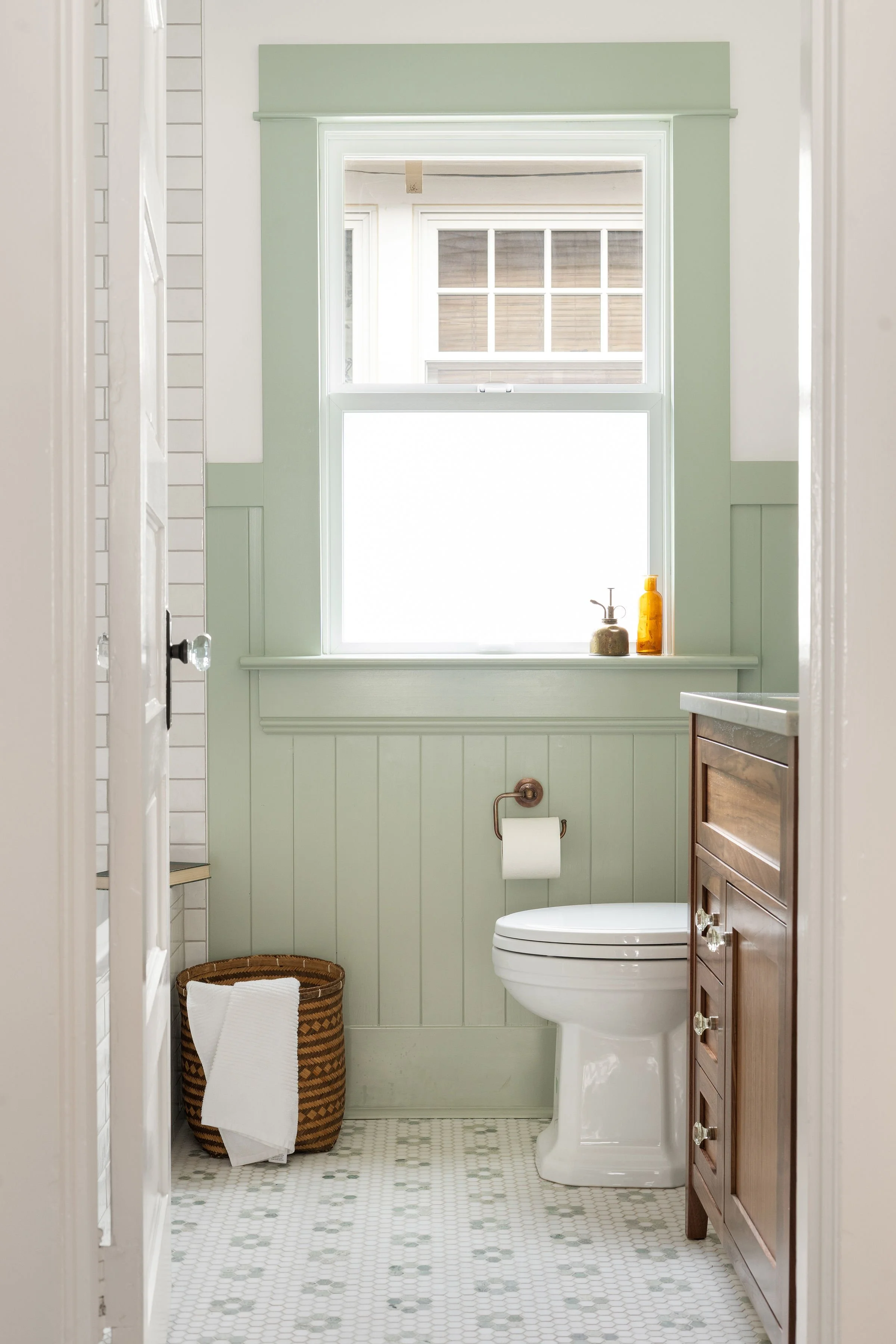 A small bathroom with a window, a white toilet, a wooden cabinet, a woven basket, and a green painted wall with white trim.