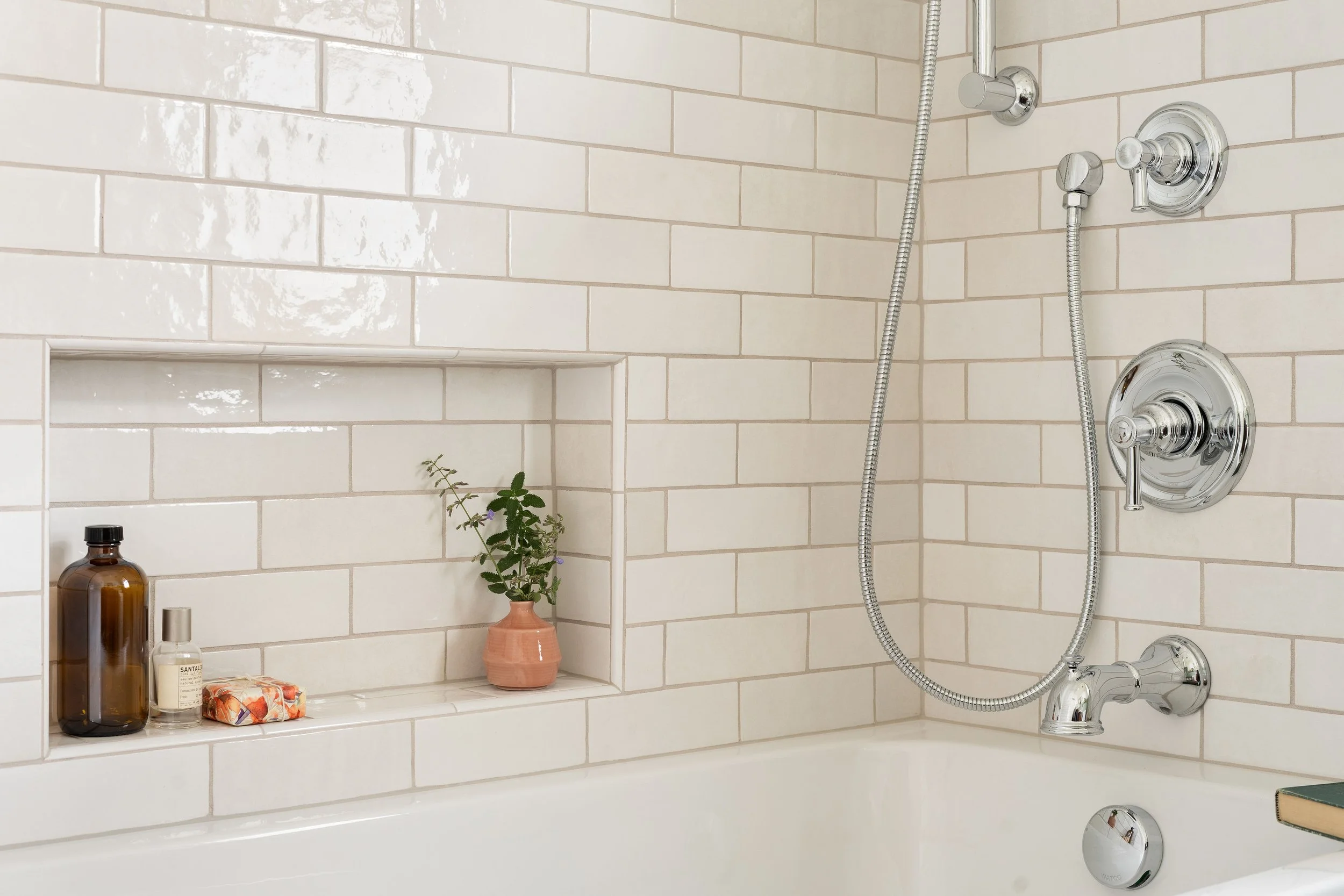 A bathtub with white subway tile walls, built-in shelf holding bottles, a small vase with a plant, and silver shower fixtures with a handheld showerhead.