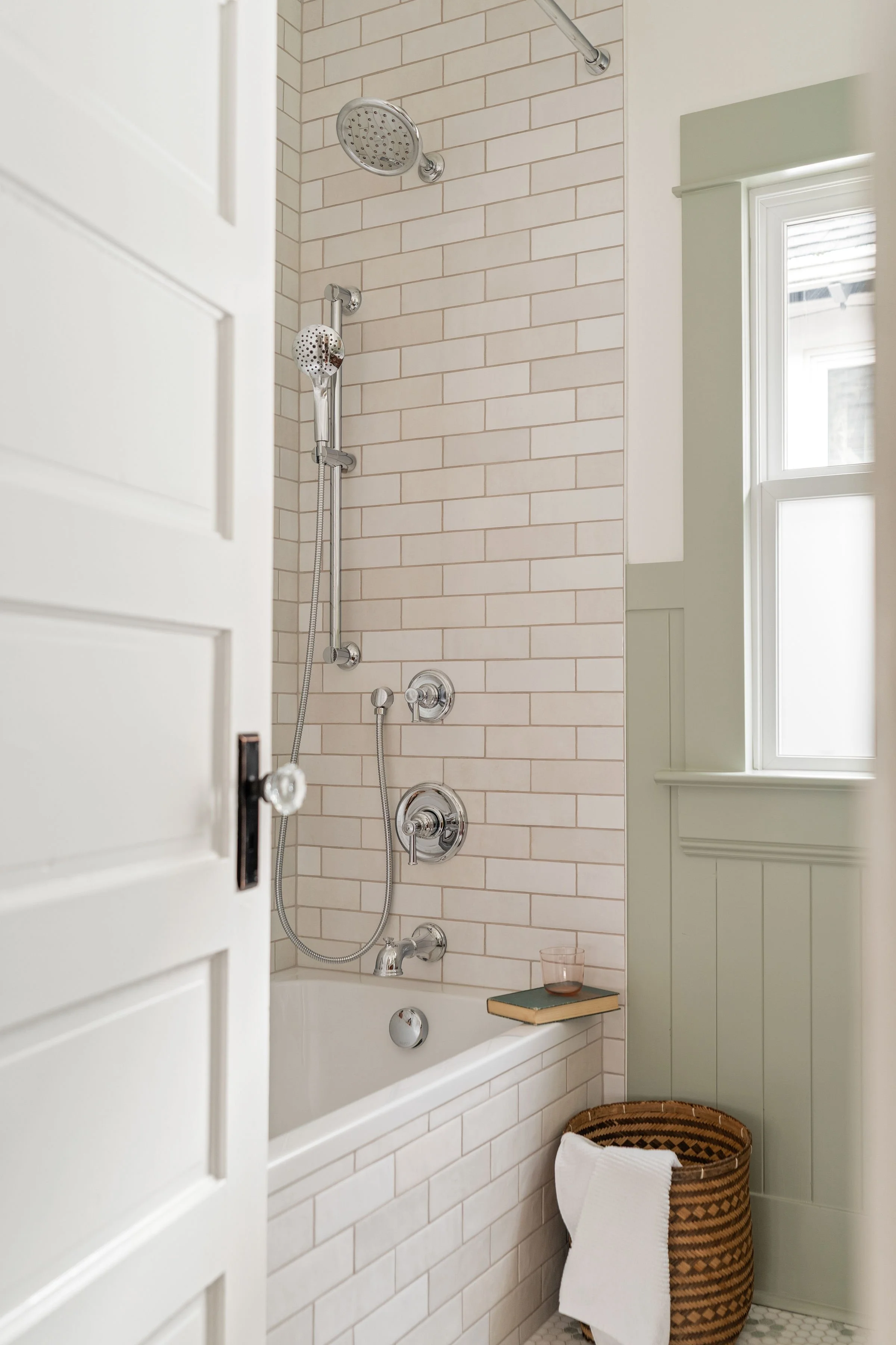 A bathroom with a bathtub, a mountain-shaped showerhead, a handheld shower, and a window beside a wainscoted wall, with a basket, towel, and books on the tub edge.