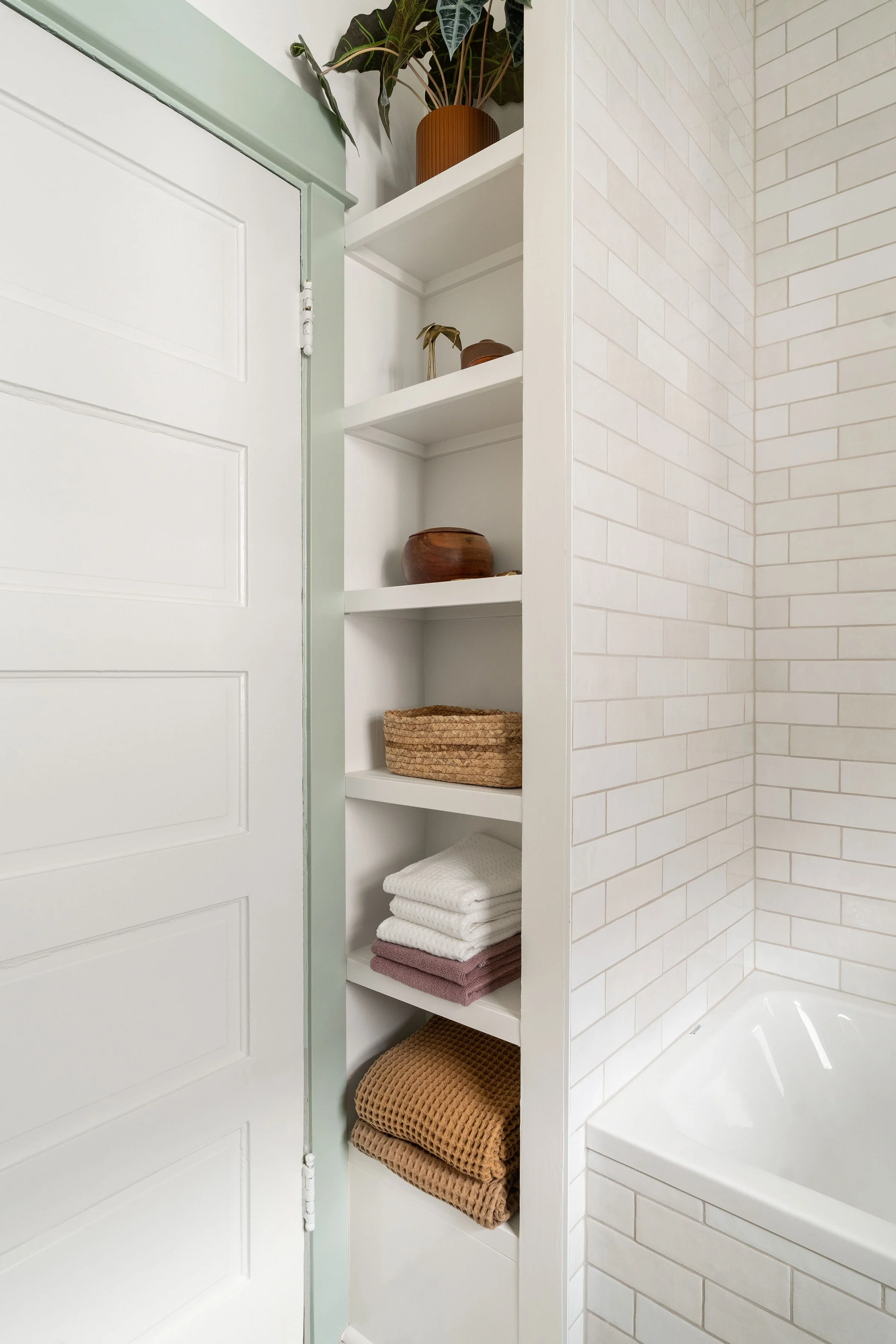 Bathroom with built-in shelves containing various towels, baskets, and decorative items, adjacent to a bathtub with white subway tile walls.