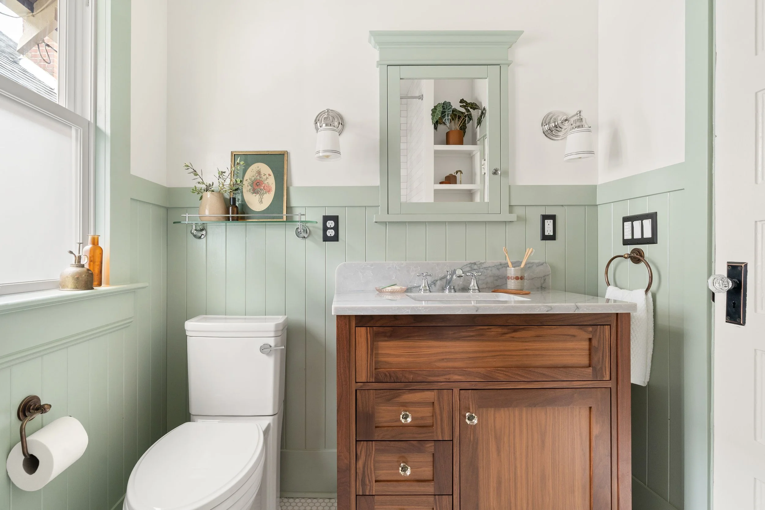 Bathroom with a white toilet, a wooden vanity with a marble countertop, a mirror cabinet, and decorative items like vases and plants.