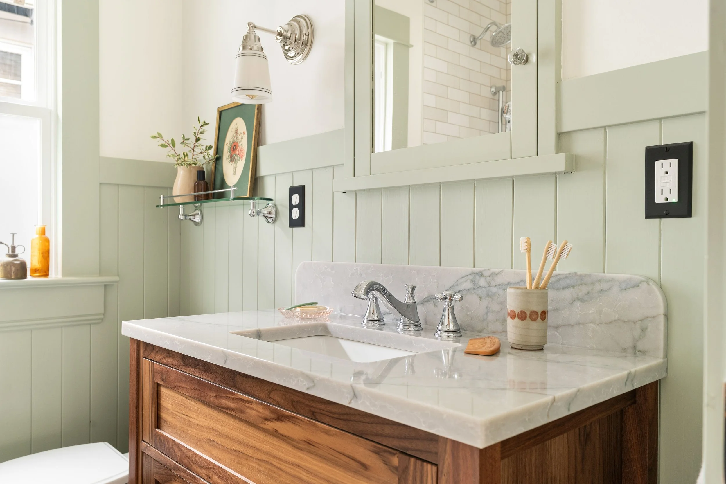 Bathroom vanity with marble countertop, wooden cabinet, and mirror. Items include toothbrushes, a soap dish, a small plate, and decorative objects on shelves.