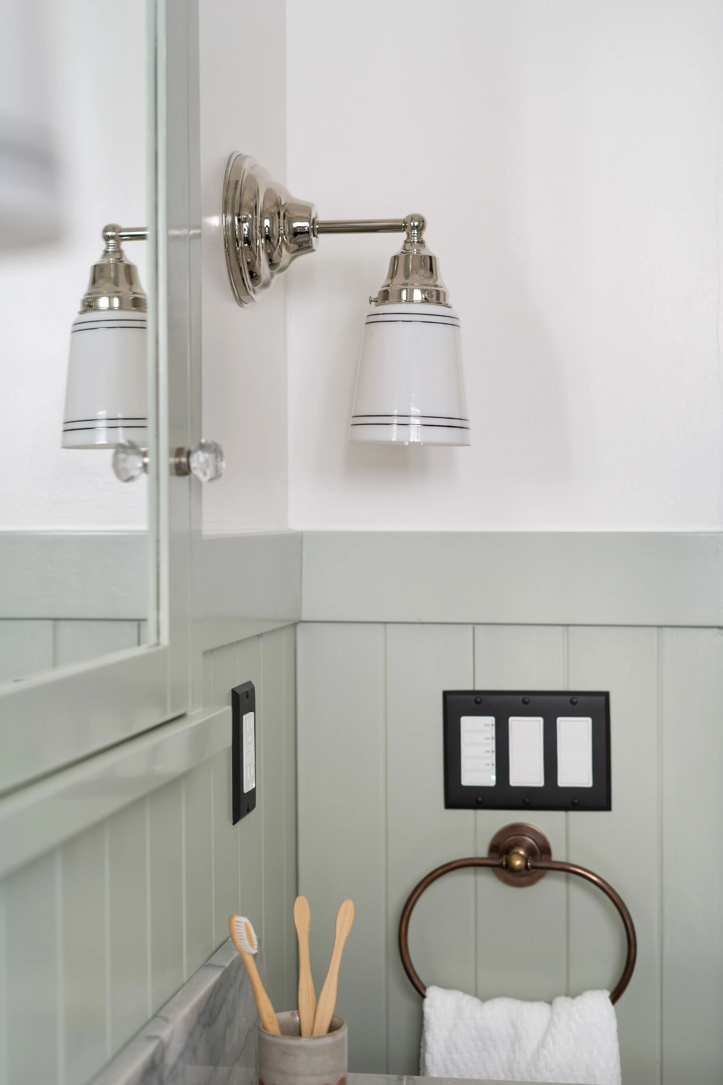 A bathroom with a white wall, a wall-mounted light fixture with two lights, a mirror, a light switch panel, and a towel ring with a white towel, holding two toothbrushes in a holder.