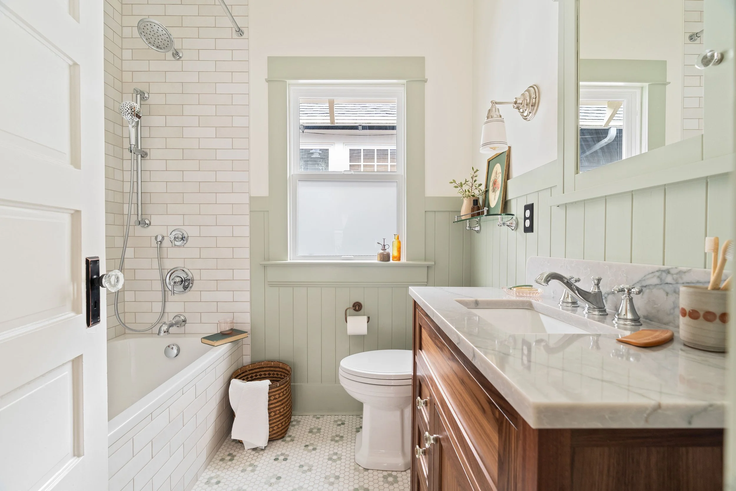 Small bathroom with bathtub, shower, window, toilet, and wooden vanity with marble countertop.