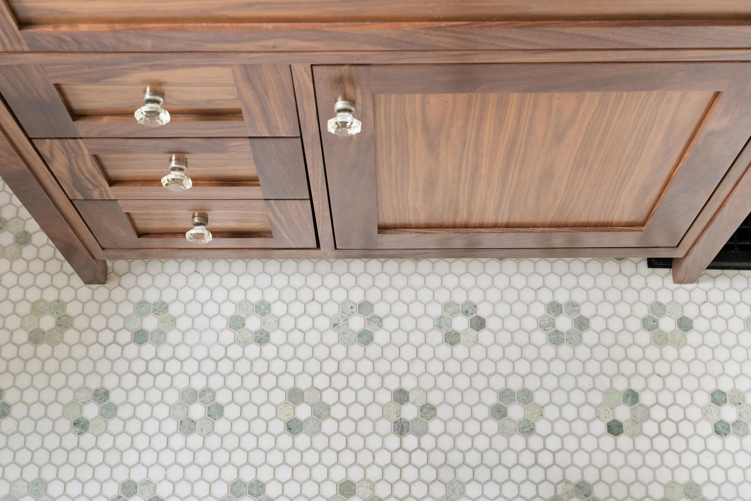 Top-down view of a wooden kitchen cabinet with glass knobs on white and green hexagon tiled floor.