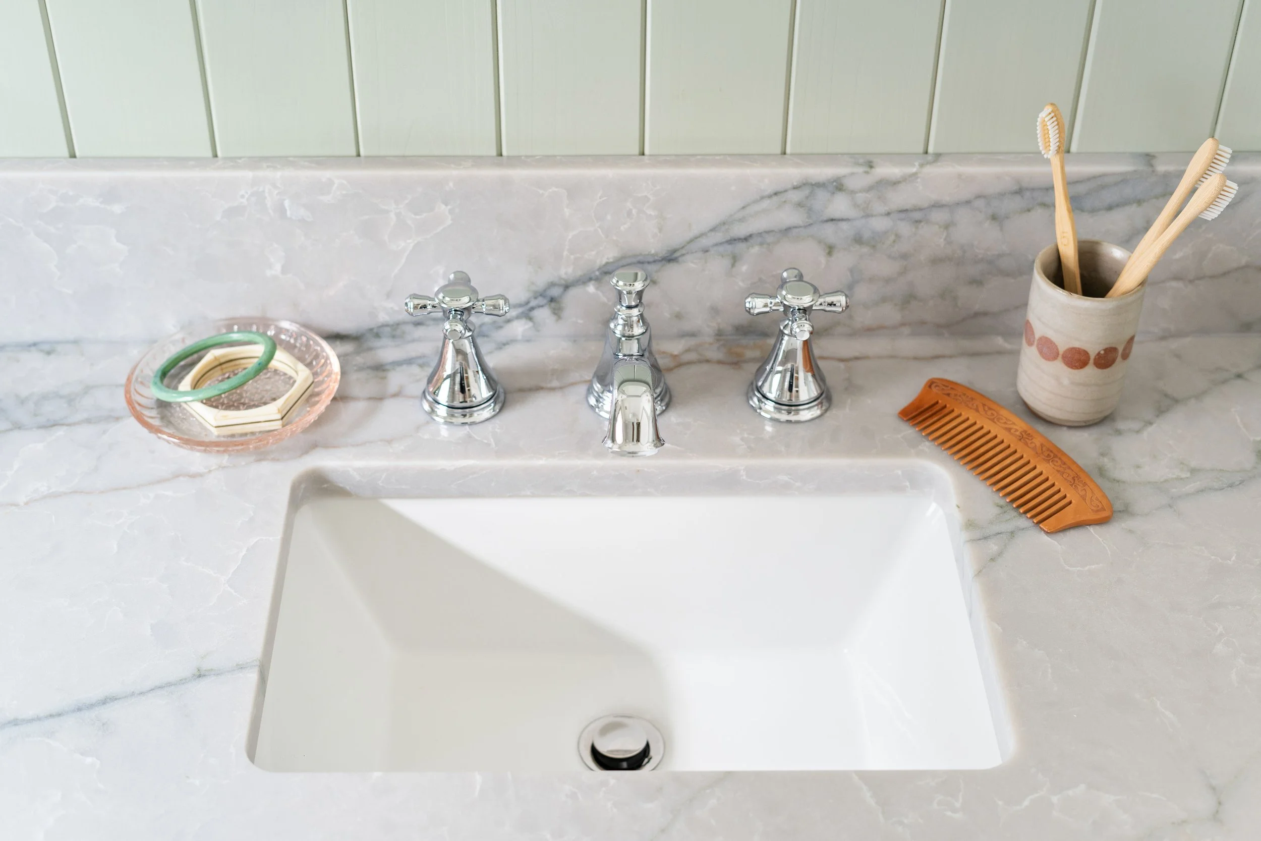 A bathroom sink with three chrome faucet handles, a marble countertop, toothbrushes in a ceramic holder, a hair comb, and decorative rings on a soap dish.