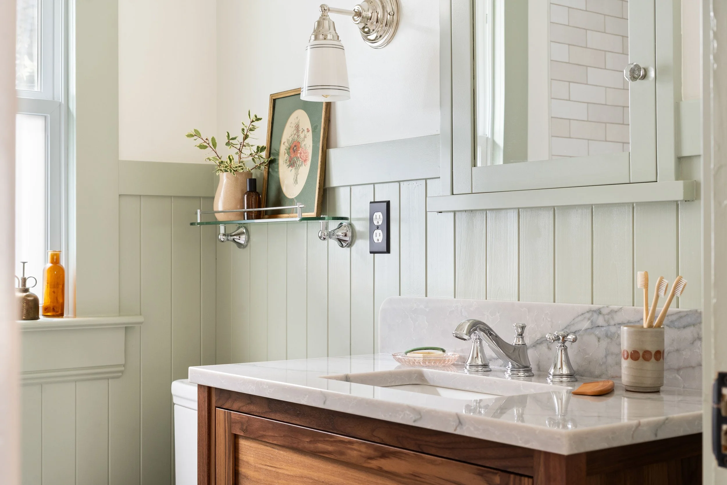 Bathroom with light green wainscoting, a white marble countertop with a sink, toothbrush holder with toothbrushes, mirror cabinet, wall sconce, framed artwork, and decorative bottles on window sill.
