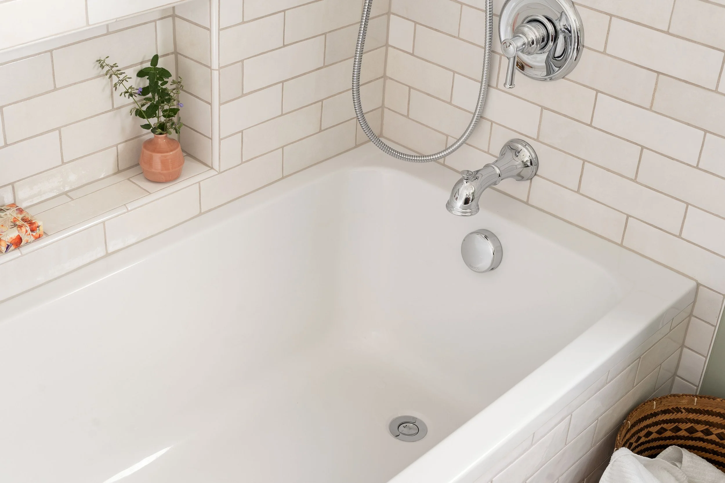 Bathroom bathtub with chrome fixtures, a showerhead with hose, a small potted plant in a pink pot, and some toiletries, with tiled walls in a neutral color.