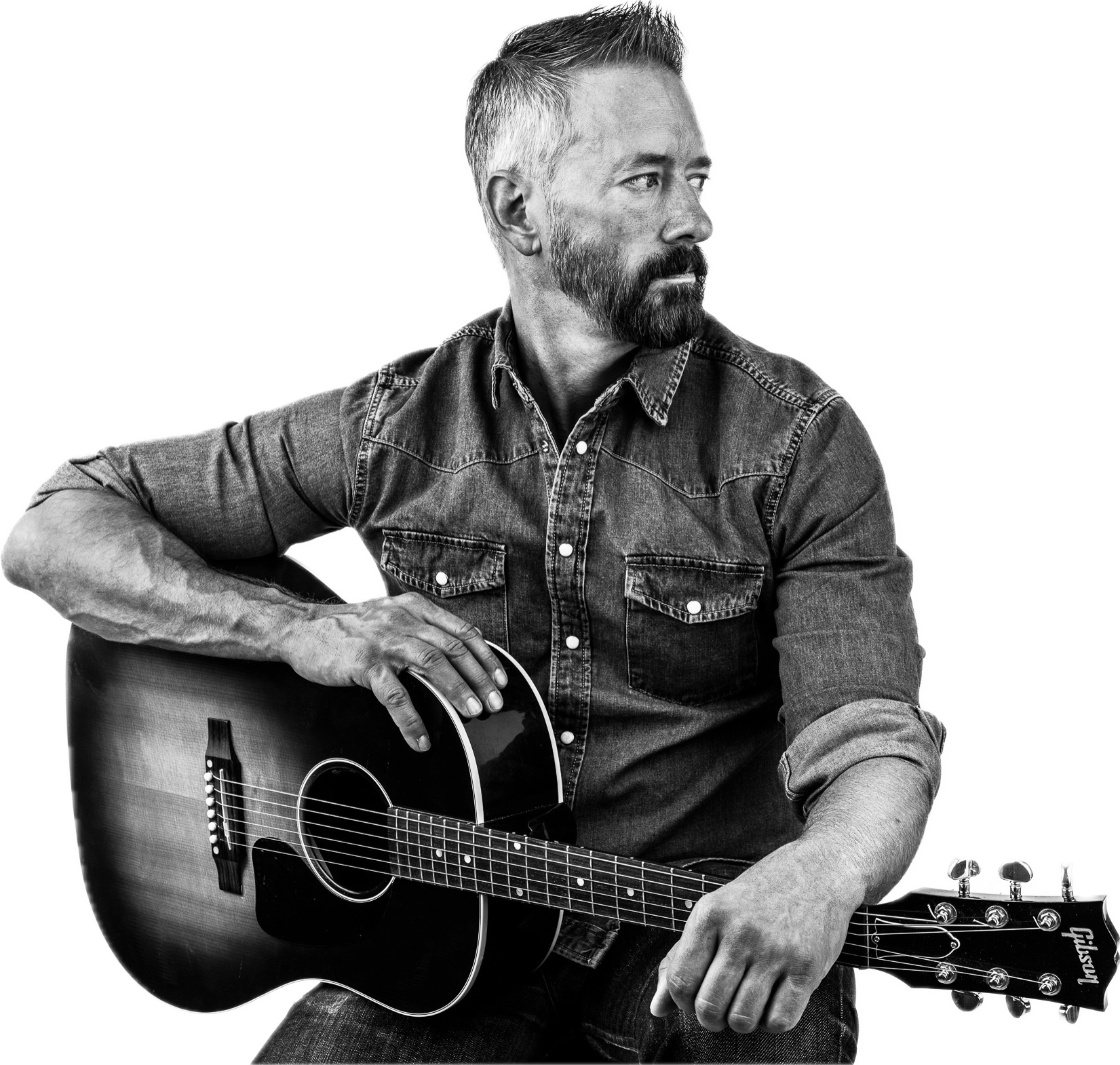 A black-and-white photo of a man with a beard and short hair, wearing a denim shirt, sitting with a guitar. The man is looking to his left and the background is plain white. He plays Americana, folk, bluegrass, and red dirt country music.