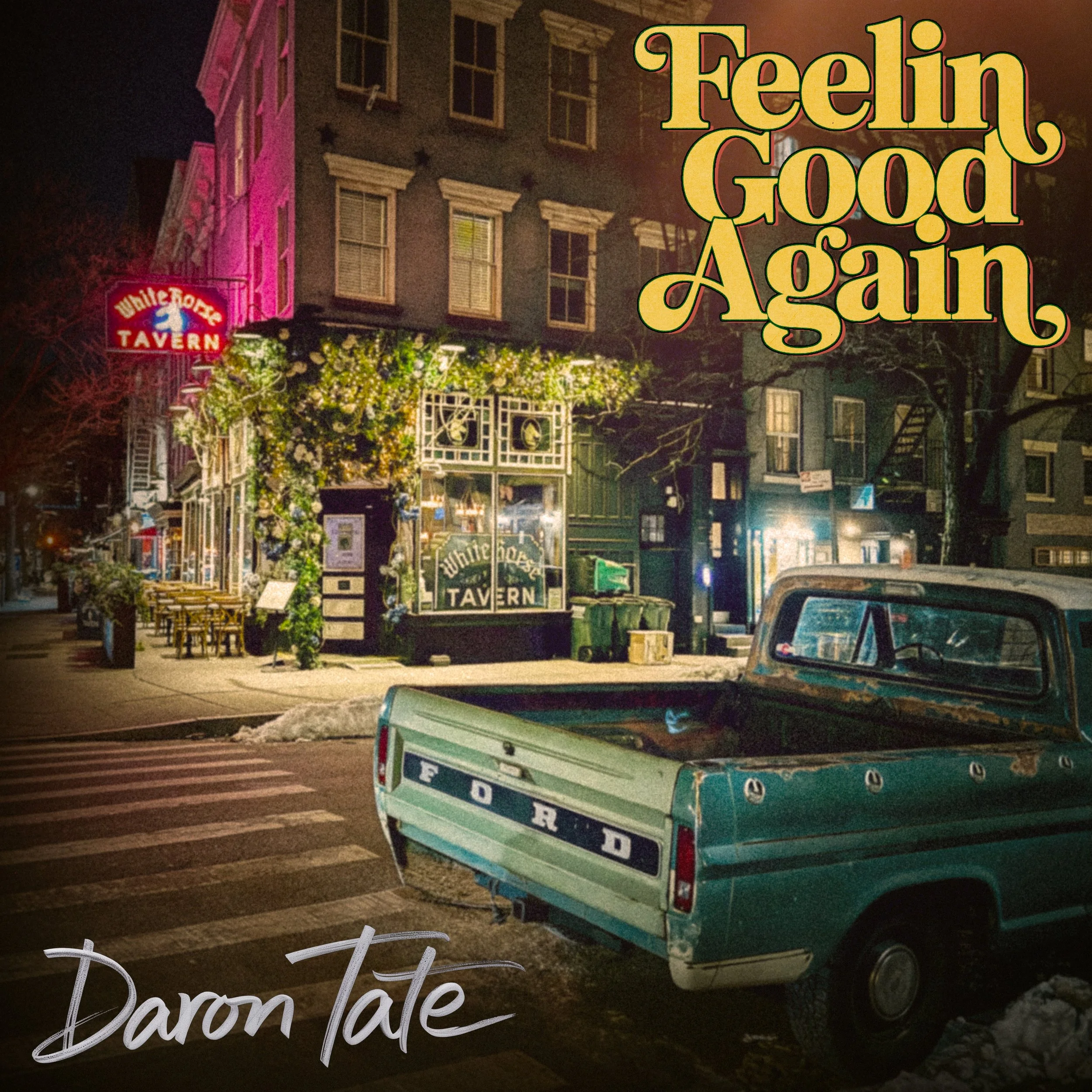 Nighttime street scene showing a multistory building with decorative window frames and a neon sign for a tavern. In the foreground, a vintage green and blue pickup truck is parked, and there are illuminated flowers and outdoor furniture along the sidewalk.