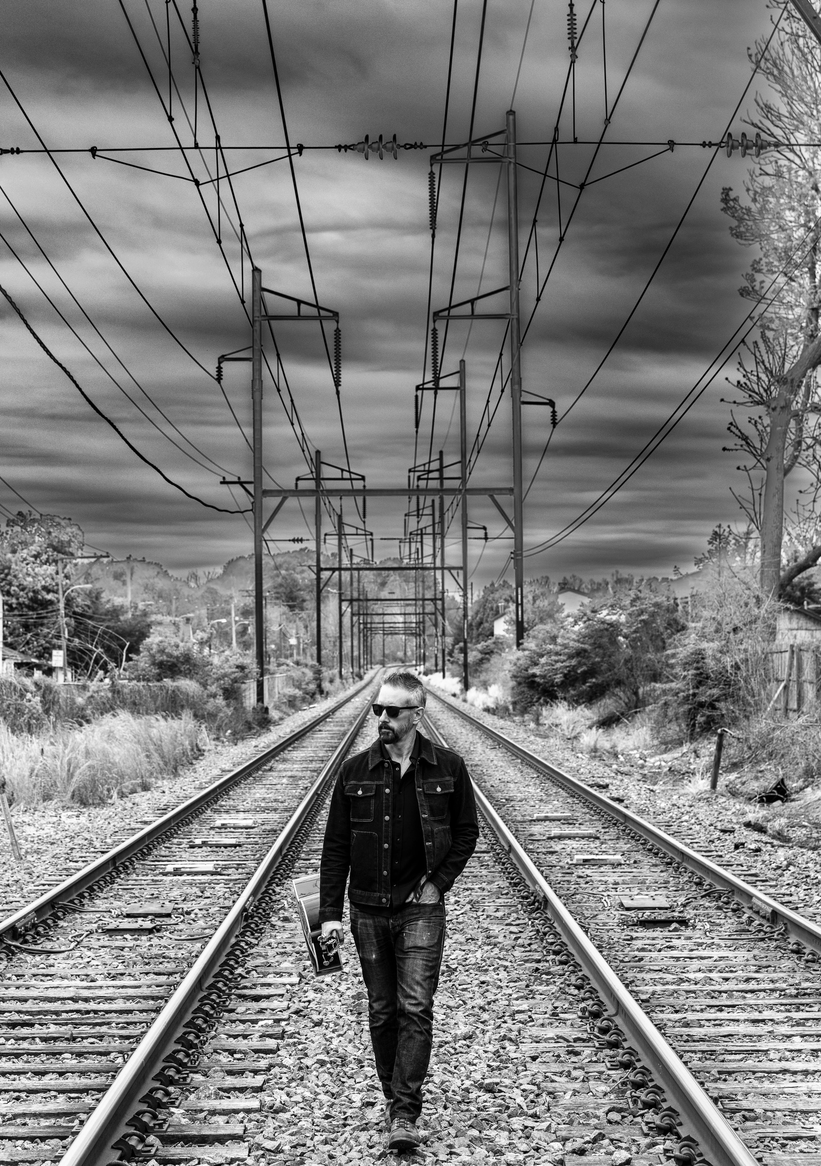 A man walking along railroad tracks holding a guitar, wearing sunglasses and a denim jacket jacket, with a cloudy sky overhead and trees on the sides.